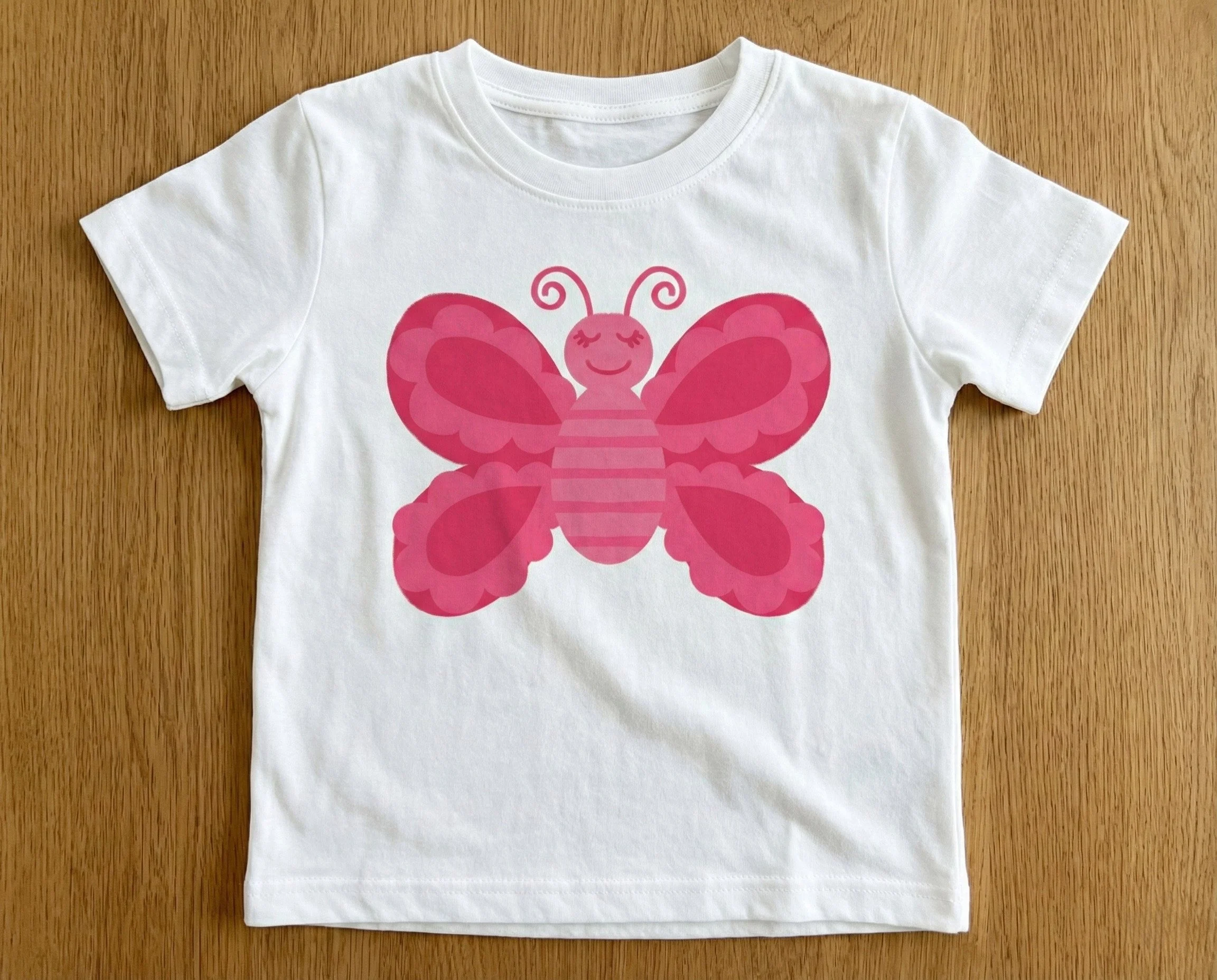 little girls white tee shirt with spot illustration of friendly cartoom butterfly in shades of pink, designed byhazel and henrietta for art licensing