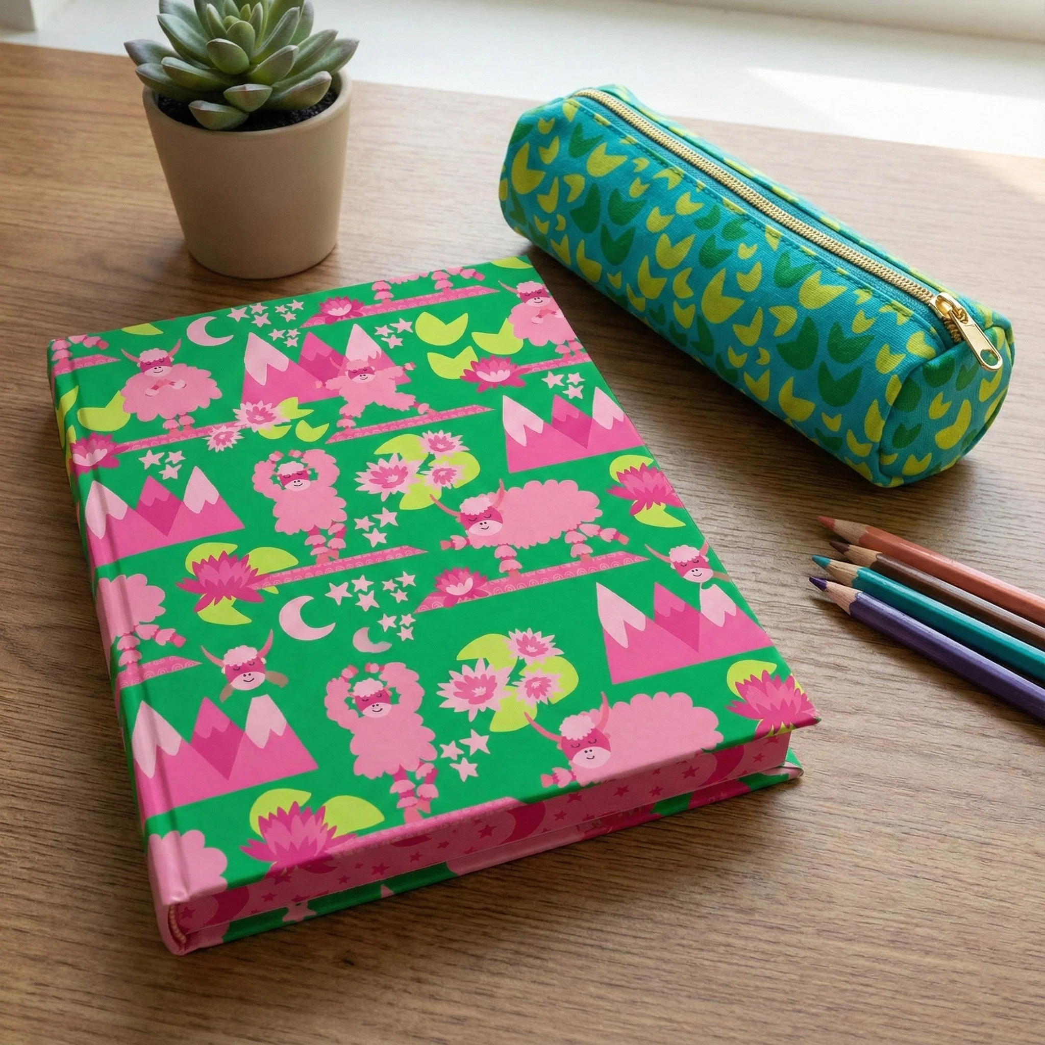 Notebook and pencil pouch featuring Yak-a-Doodle Yoga surface pattern designs with playful yaks, lotus flowers, lily pads, mountains, moons, and stars in bright green, pink, and turquoise.Yak-a-Doodle Yoga surface pattern designs shown on stationery 