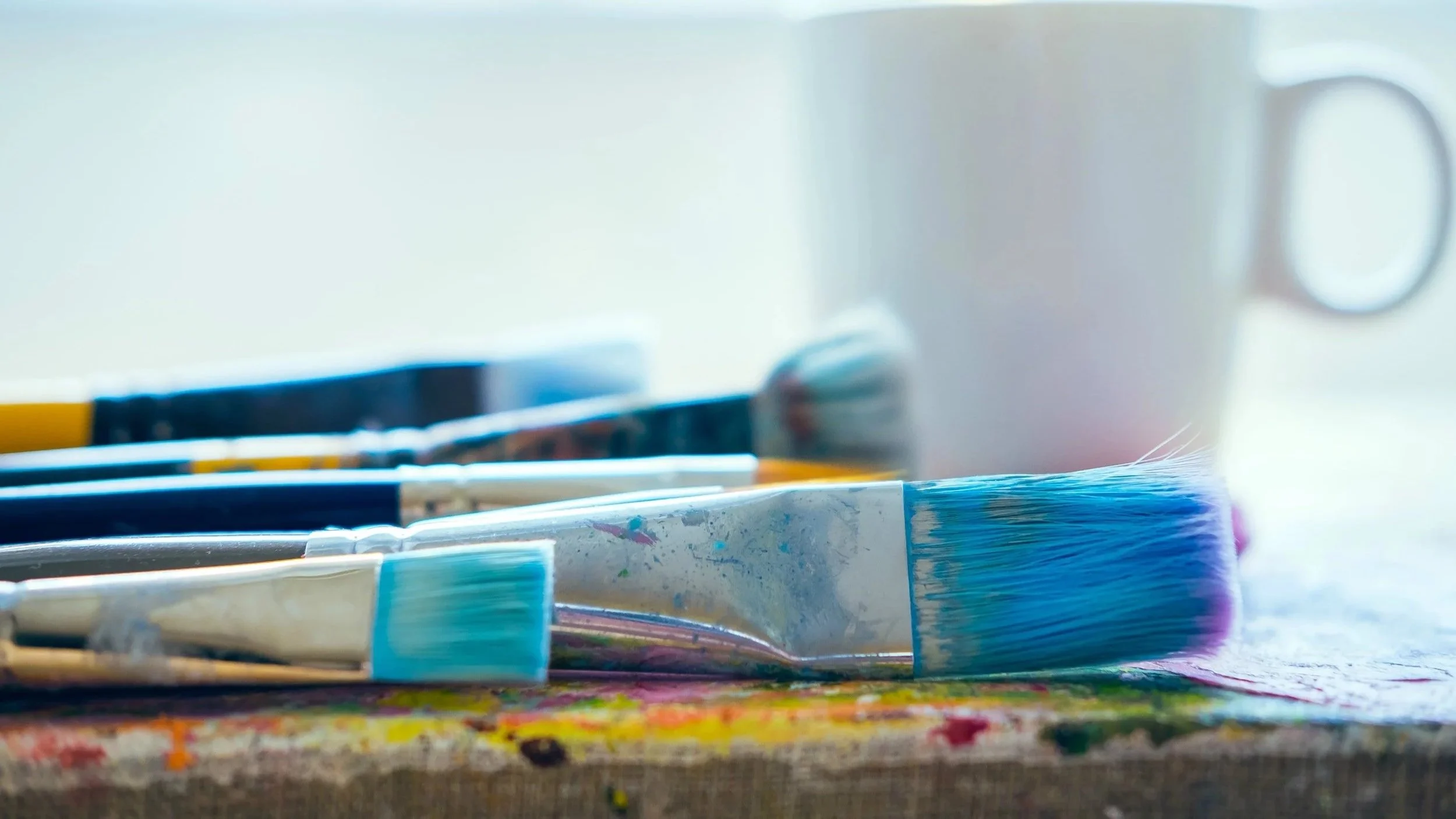 Close-up view of several paintbrushes with colorful paint on a surface, and a white mug in the background.