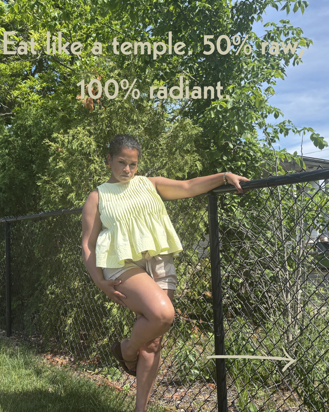 Keenya Willis posing against a black chain link fence in a yellow top with text overlay reading Eat like a temple 50 percent raw 100 percent radiant representing intentional nourishment for midlife women