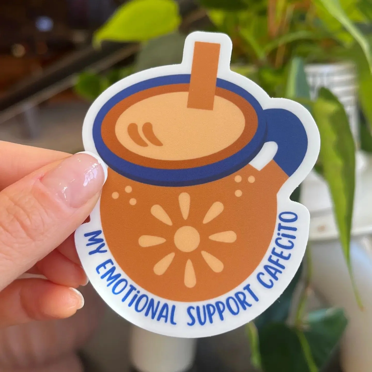 Hand holding a sticker of a coffee cup labeled "My Emotional Support Cafecito"
