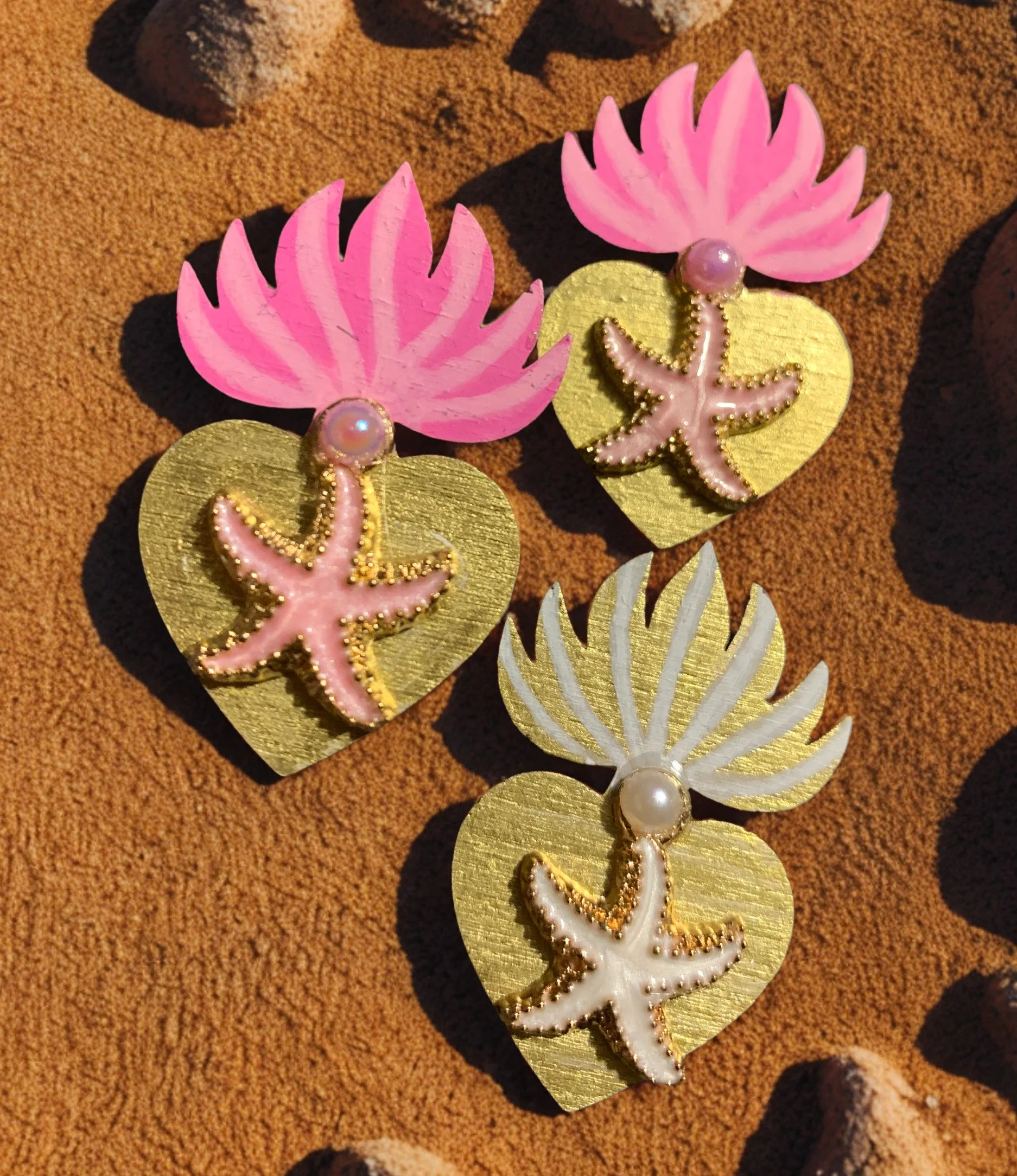 Three pink, white and gold sea star pins on sandy beach