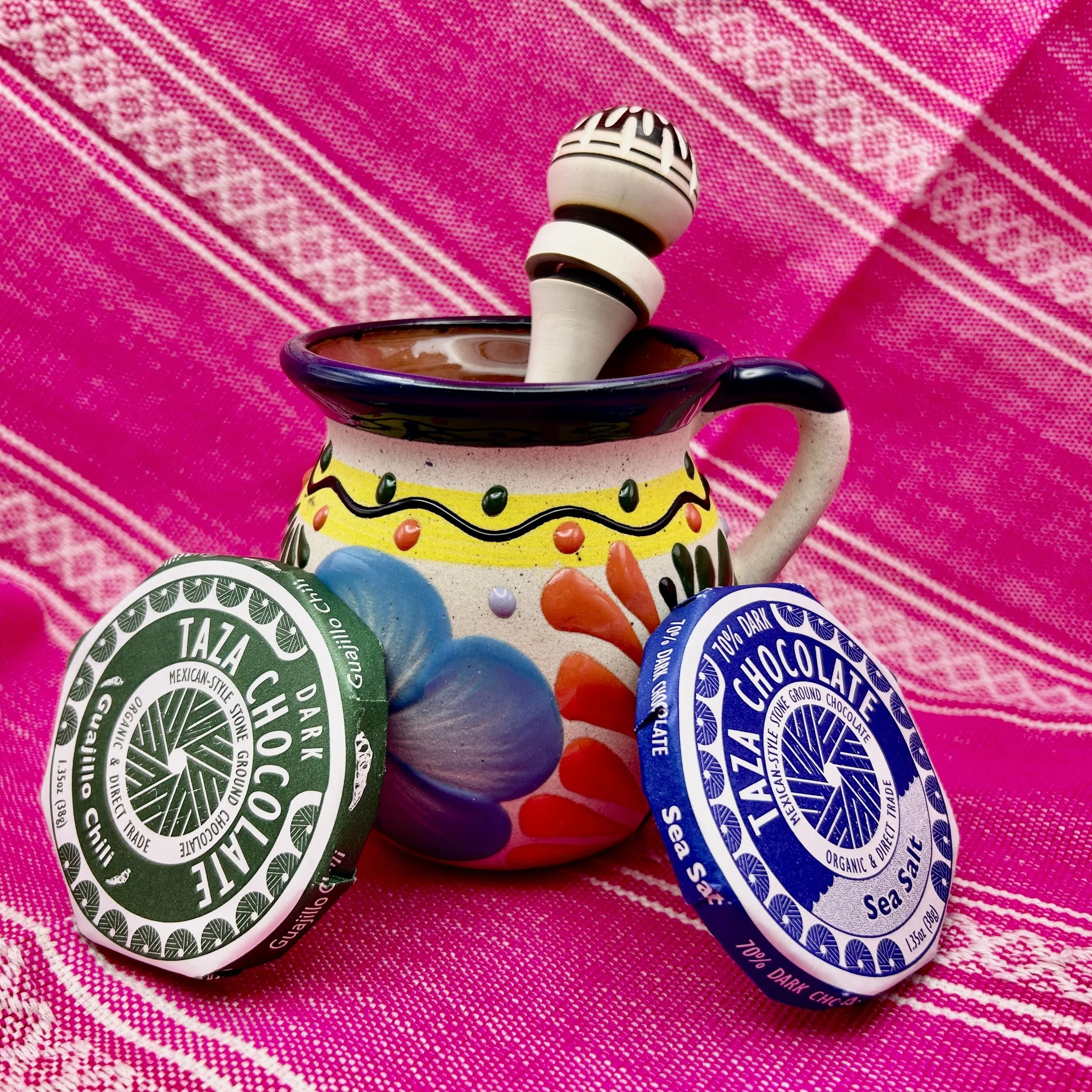 Colorful decorative mug with wooden frother, Mexican-style Taza chocolate discs on vibrant pink textile background.