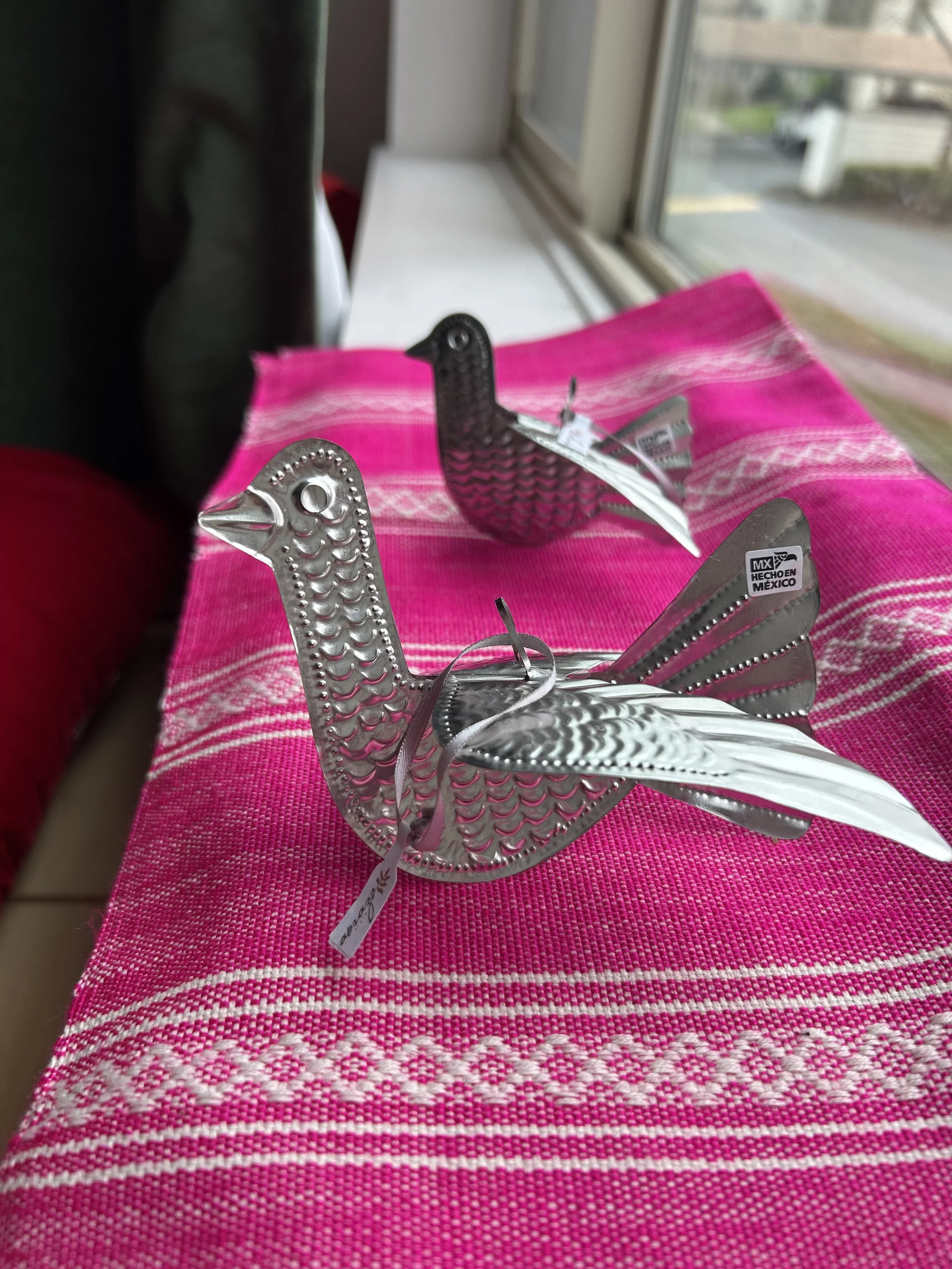 Two metallic bird sculptures on a pink woven fabric placed near a window with an outdoor view.