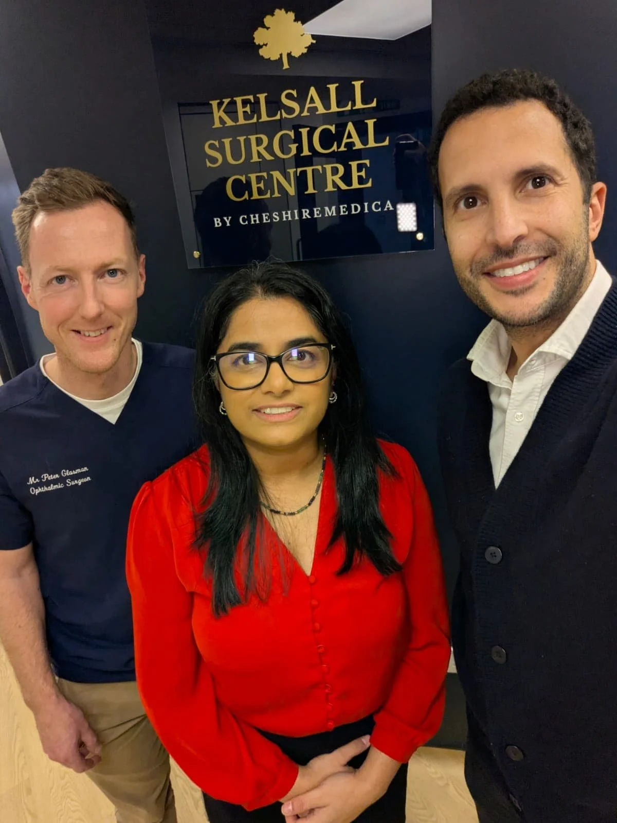 Three people taking a selfie in front of a sign that reads 'Kelsall Surgical Centre by Cheshire Medical'. The person in the middle is a woman with black hair, glasses, wearing a red blouse. The person on the left is a man with light skin, short reddish hair, wearing a navy scrub top with embroidered text, and the person on the right is a man with light skin, dark curly hair, wearing a white shirt and dark sweater.