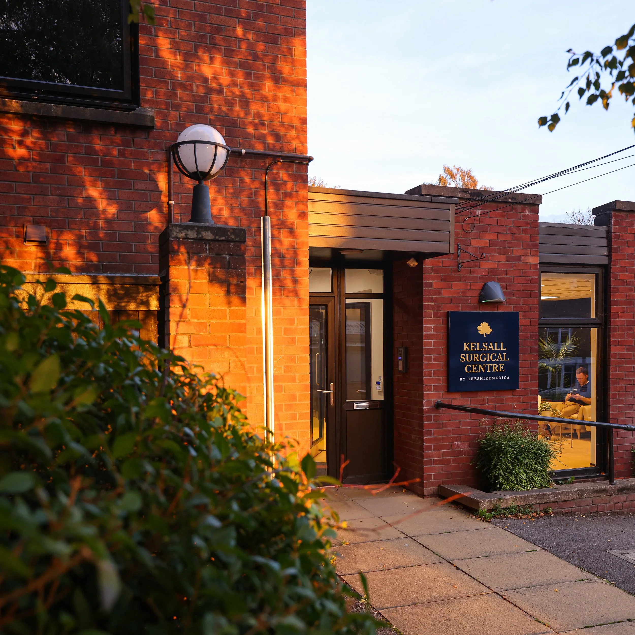 Exterior of a medical center building with a sign reading 'Kelsall Surgical Centre by Cheshire Medica.' The building is made of red brick, with a black door, a large window showing a person sitting inside, and outdoor lighting fixtures. The scene is illuminated by warm sunlight, with some greenery in the foreground.