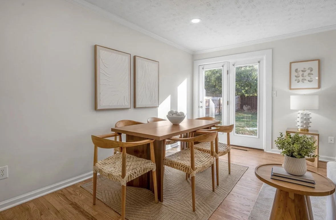 Dining room with a wooden table, six chairs, and a bowl on the table. Glass doors lead to a backyard. Wall art, a lamp, and a side table with a plant are visible.