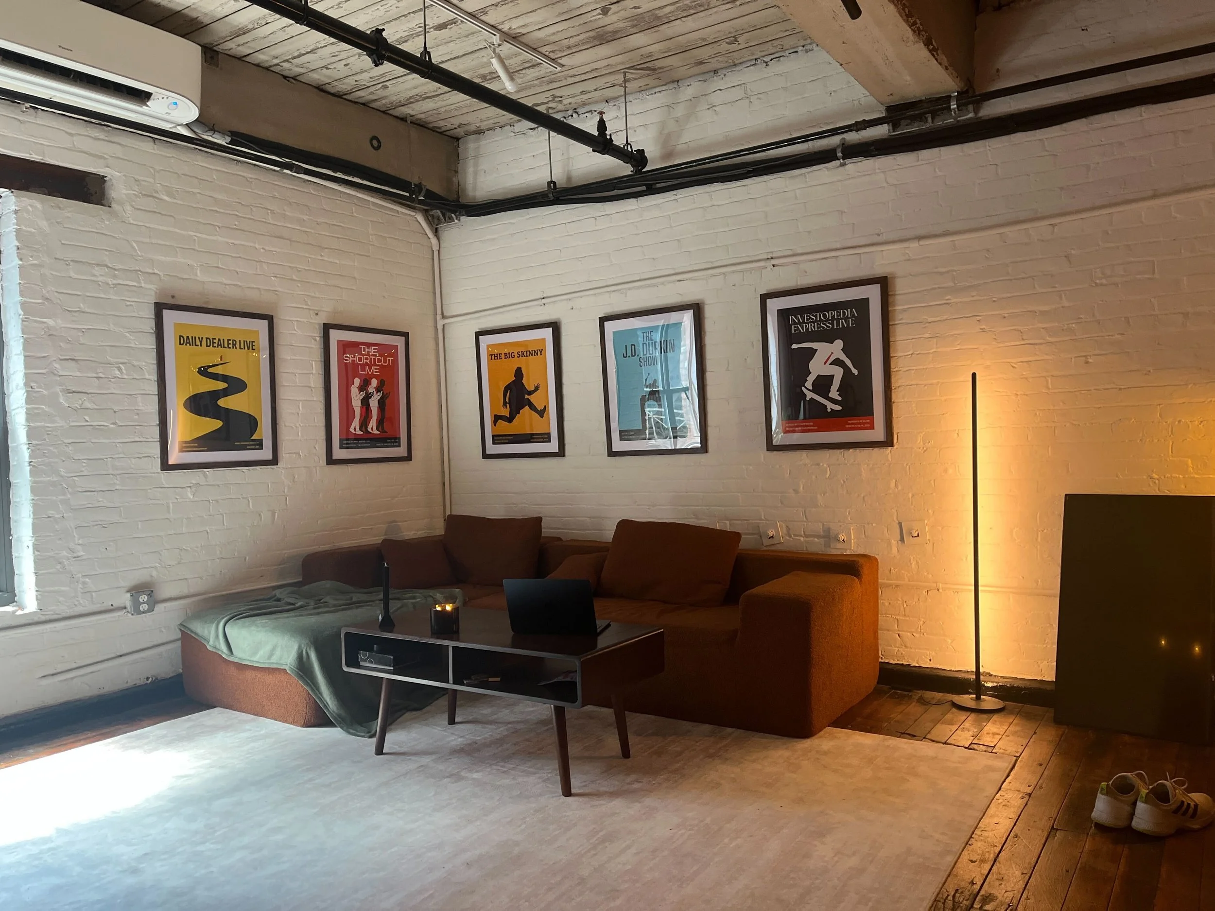A cozy living room with a white brick wall decorated with framed posters, a brown sectional sofa, a coffee table, and a floor lamp. Shoes are placed at the edge of a wooden platform near the wall, and there is an air conditioning unit mounted on the upper wall. The ceiling features exposed wooden beams and black piping.