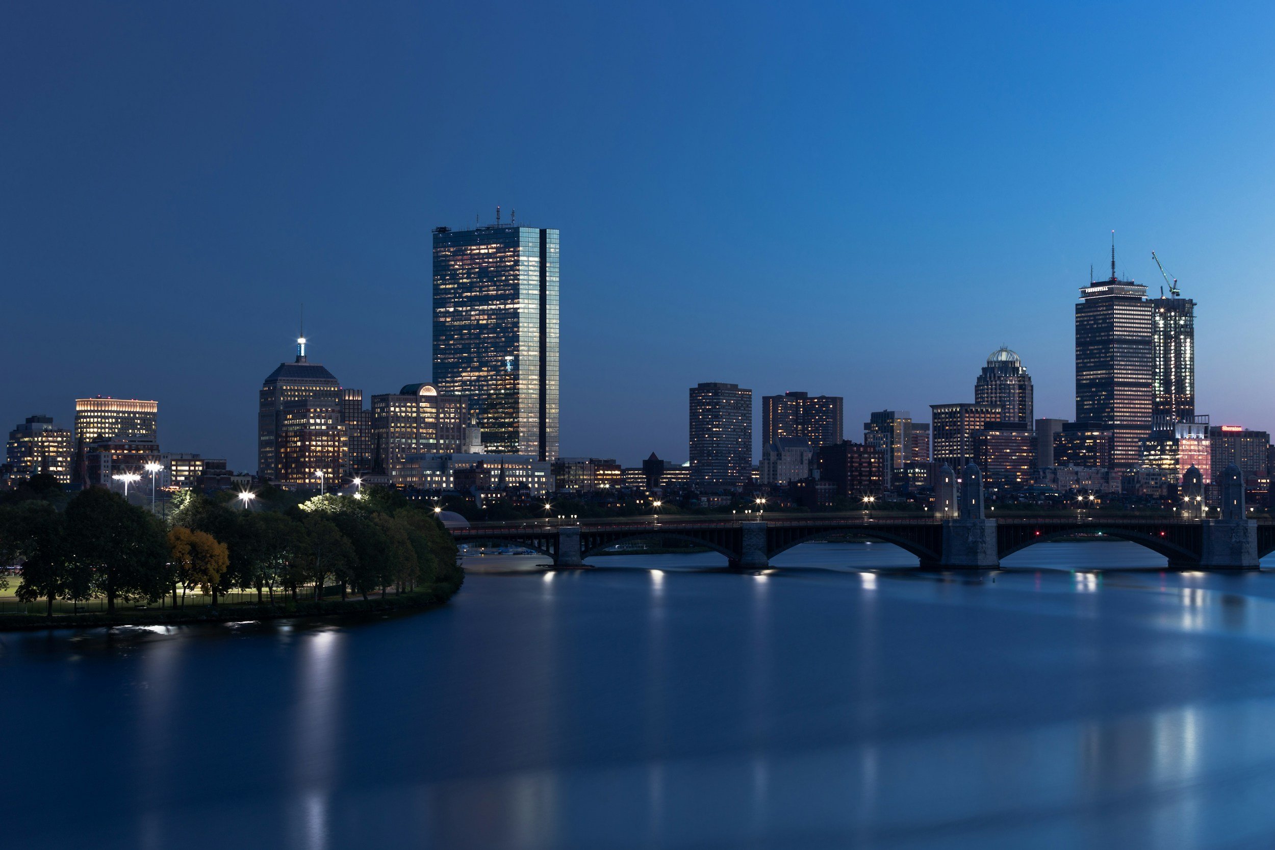Nighttime city skyline with tall modern buildings reflected on a river, with a bridge in the foreground and trees along the riverbank.