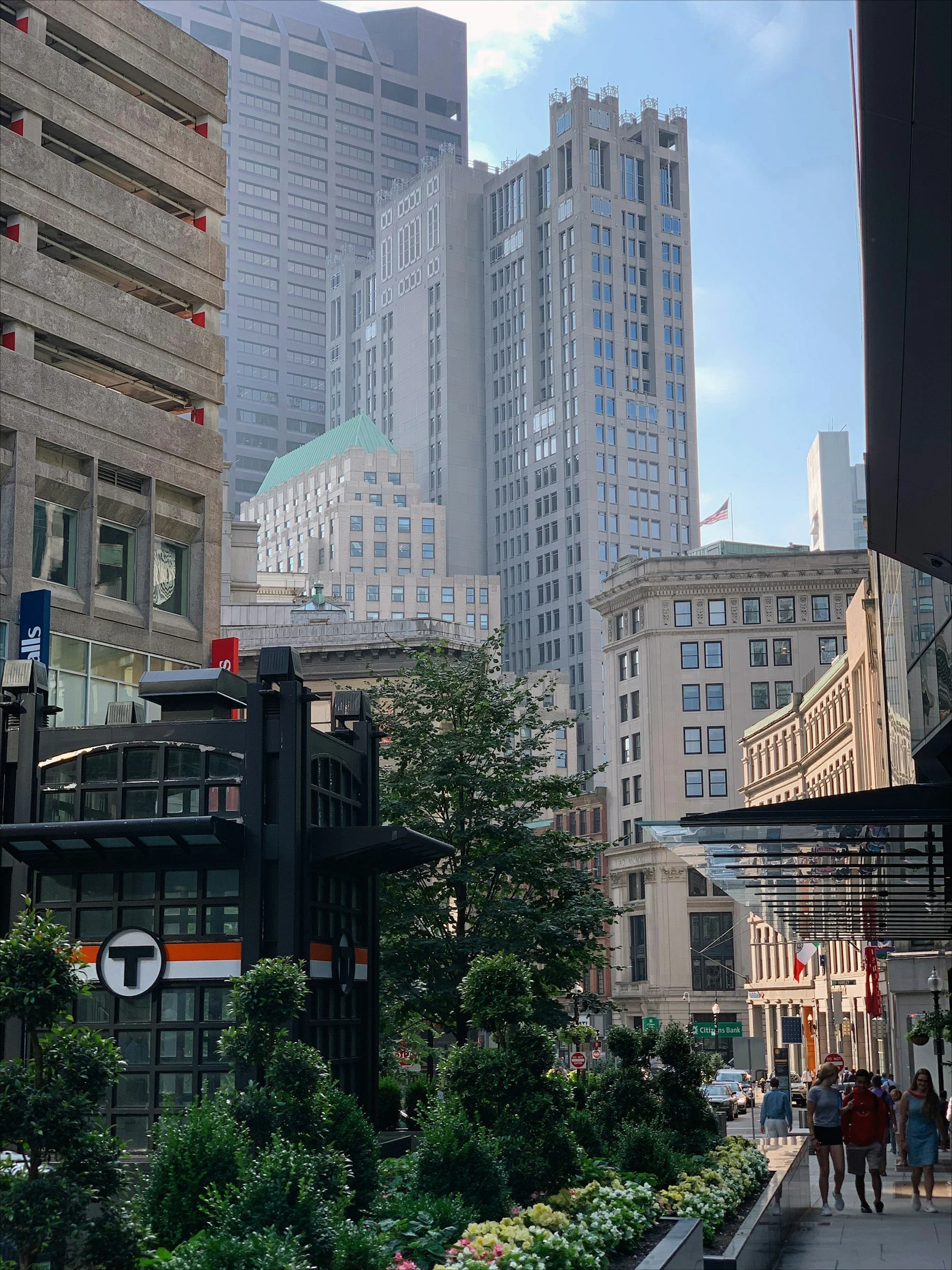 City street scene with pedestrians, tall office buildings, a subway entrance marked with a 'T' logo, and landscaped greenery along the sidewalk.
