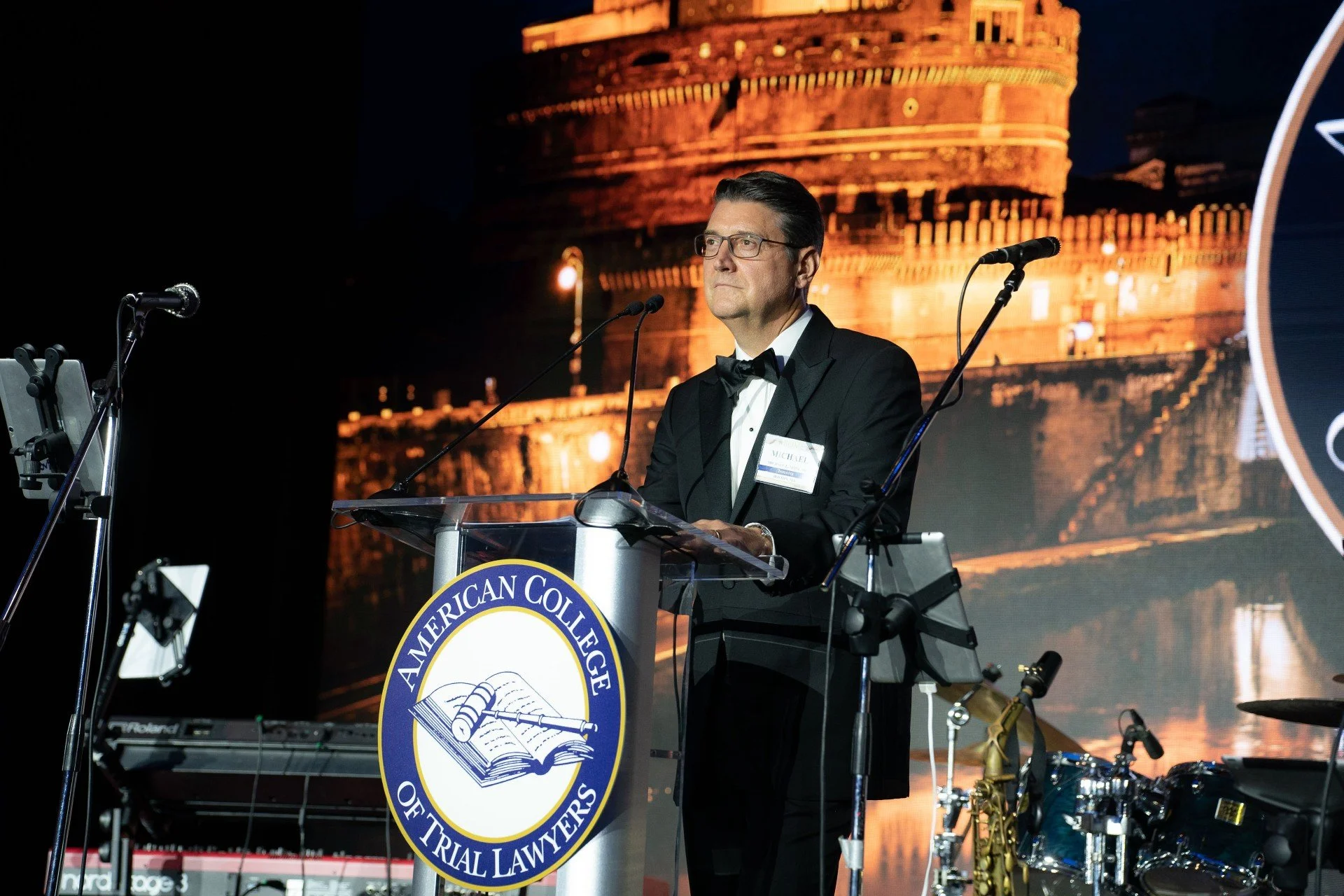 A man in a black tuxedo and glasses stands at a podium with the American College of Trial Lawyers emblem. He is giving a speech at an event, with a large image of a historical building illuminated at night in the background. There are musical instruments and microphones around him.