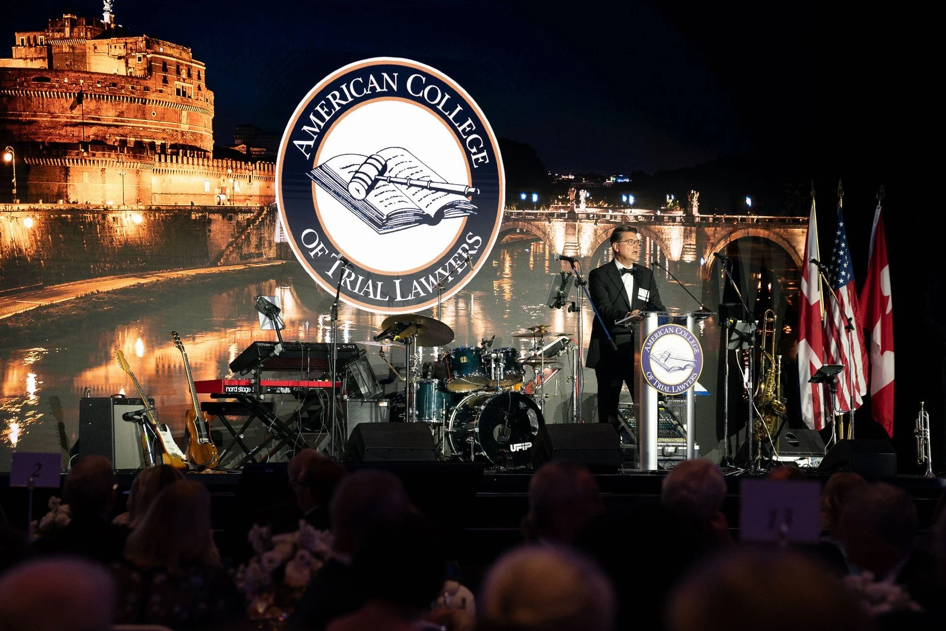 A man in a tuxedo speaking at a podium with a large backdrop displaying the logo of the American College of Trial Lawyers, with flags nearby, during an event or ceremony at night.