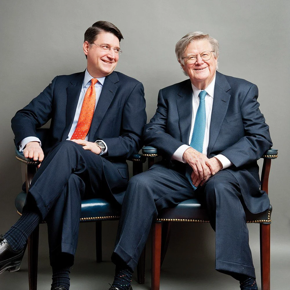 Two men in suits sitting on chairs, smiling and looking at each other against a plain gray background.