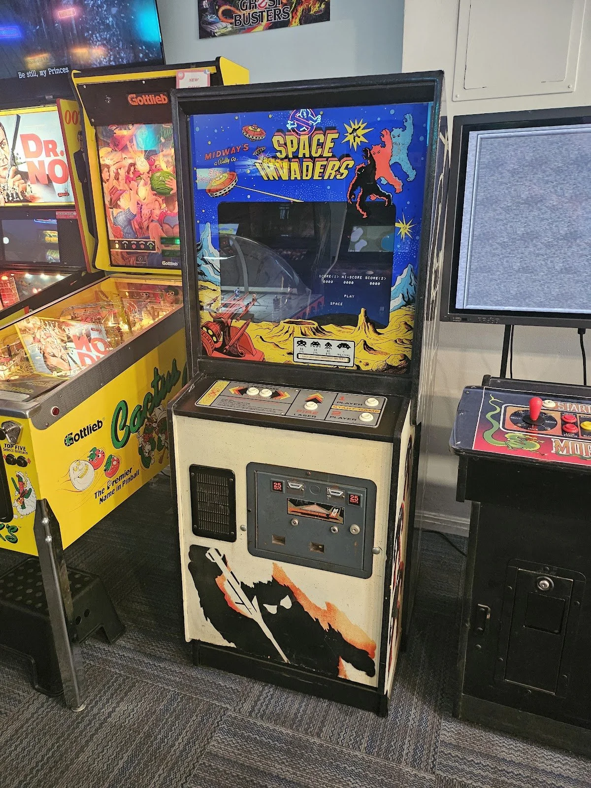 Vintage space invaders arcade game with a painted black silhouette of a soldier holding a rifle on the lower side of the cabinet and colorful space-themed artwork on the screen's frame, situated in an arcade room alongside other pinball and arcade machines.