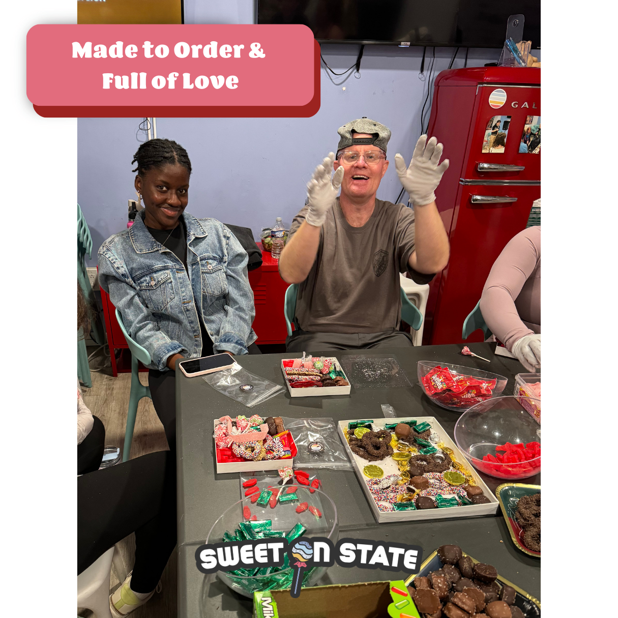 Two people sitting at a table with colorful candies and chocolates, one smiling at the camera and the other gesturing with gloved hands, in a room with a red refrigerator and a wall-mounted TV, celebrating with a sign that reads 'Made to Order & Full of Love' and a sticker saying 'Sweet on State'.