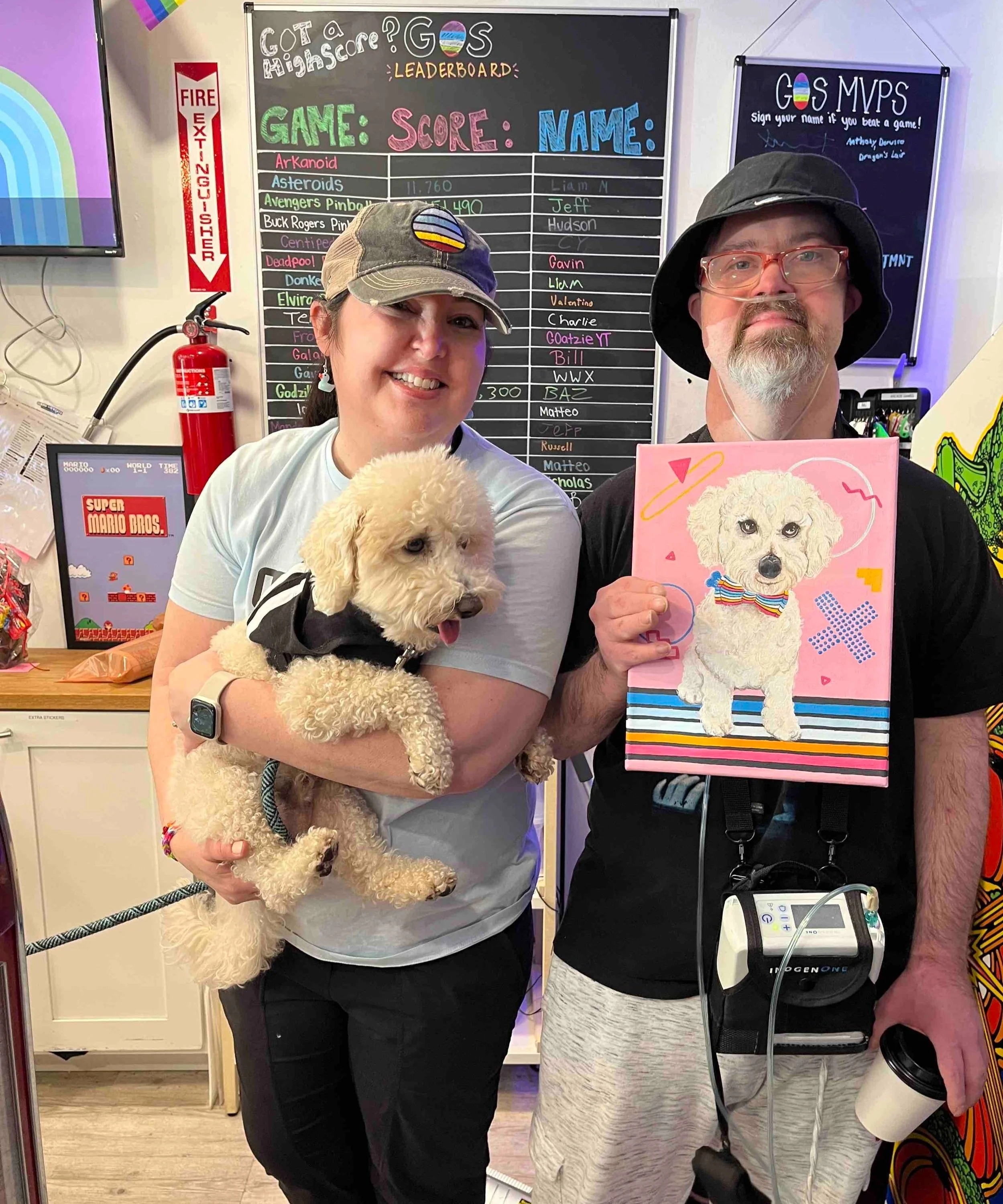 Two people posing in front of a leaderboard at a game event, one holding a fluffy dog and the other holding a colorful portrait of a dog. The woman on the left is smiling and wearing a cap, while the man on the right is holding a painting and wearing glasses and a large hat.