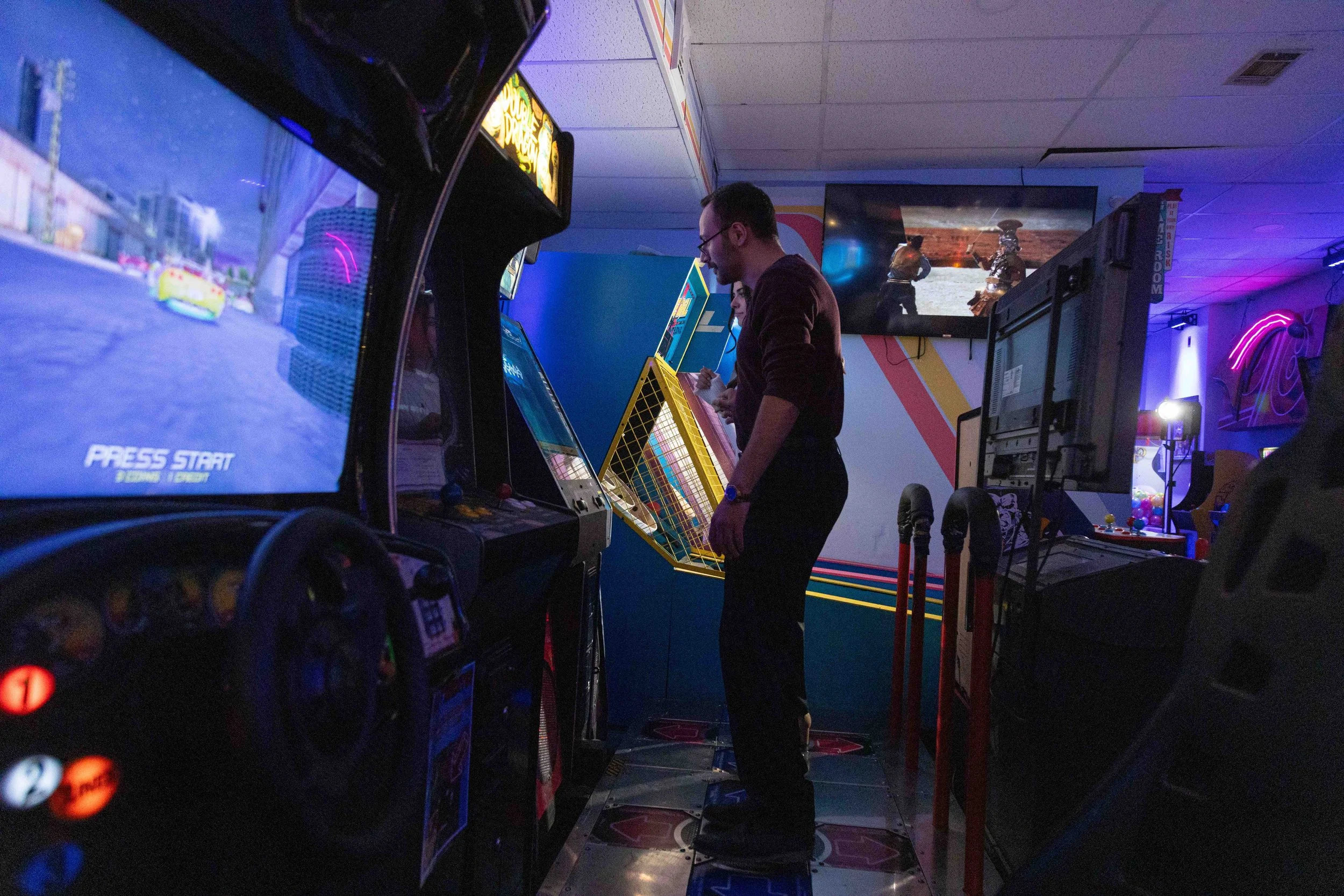 Man standing on an arcade dance game floor in a dimly lit arcade, with screens and arcade machines around.