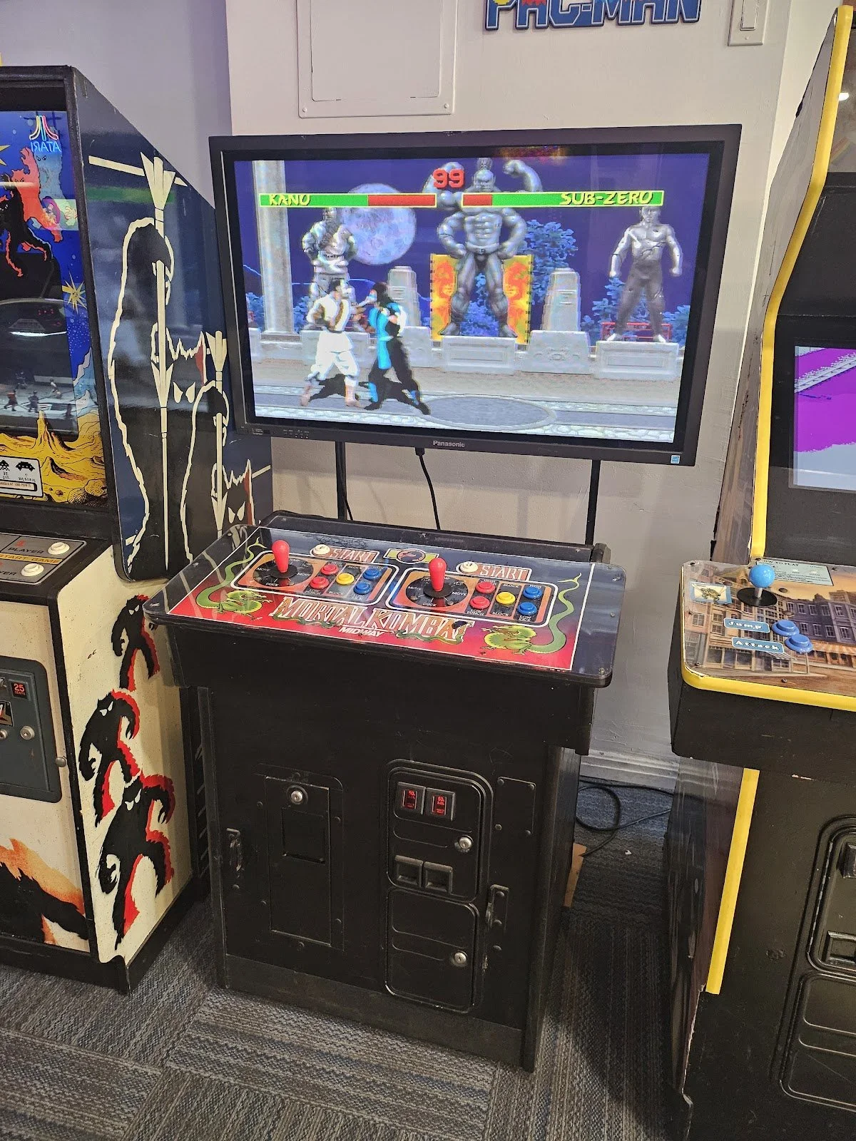 Arcade gaming setup featuring a Mortal Kombat arcade machine with a fighting game on the screen, displaying characters fighting against statues. The cabinet has a red and black control panel with joysticks and buttons, positioned next to other arcade games.