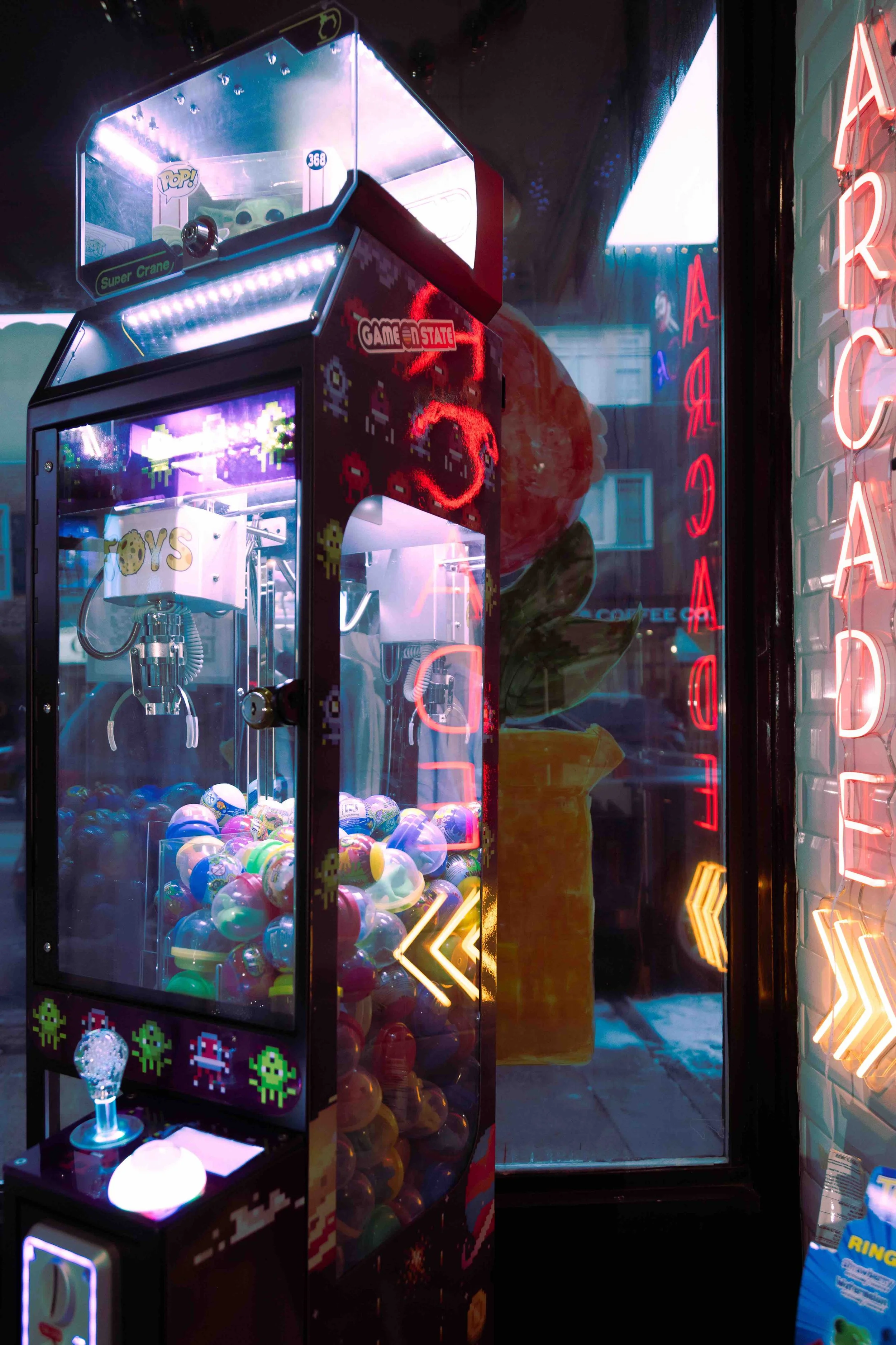 A neon-lit arcade claw machine filled with colorful toy capsules and a small toy inside, with neon signs and reflections in the background.