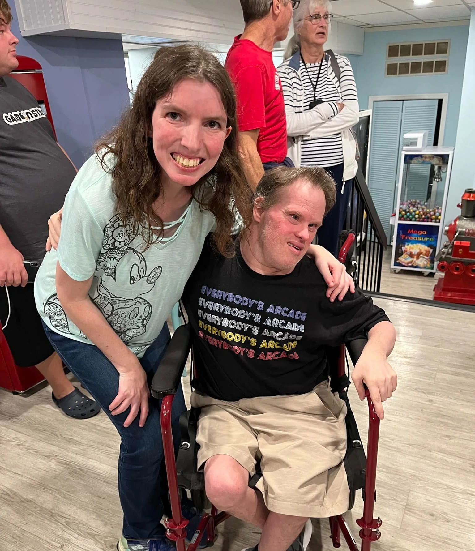 A woman with long brown hair and a big smile is leaning over and placing her hand on the shoulder of a man sitting in a motorized wheelchair. The man has short brown hair, is wearing a black T-shirt with colorful text that says 'Everybody's Arcade,' and beige shorts. In the background, there are several people in a casual setting, some talking. The room has light blue walls, arcade machines, and a wood floor.
