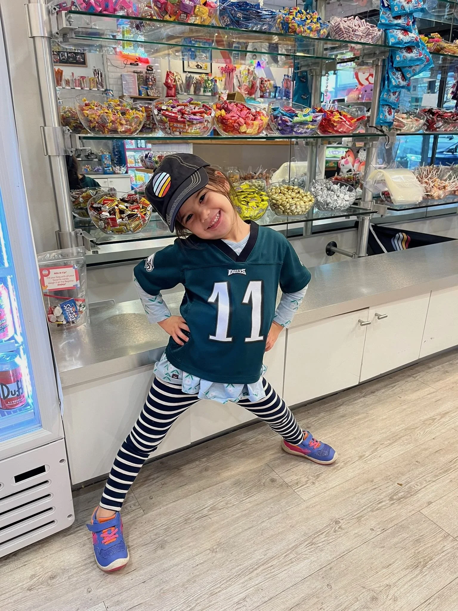 A young girl standing in front of a candy display at a store, smiling and posing with her legs apart. She is wearing a Philadelphia Eagles football jersey with the number 11, striped leggings, colorful sneakers, and a cap.