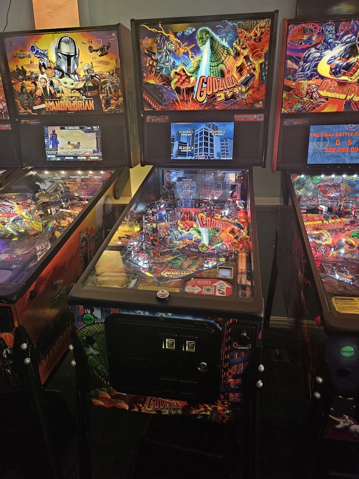 A row of vintage pinball machines featuring themed artwork from popular movies such as Godzilla, Mandalorian, and Deadpool.