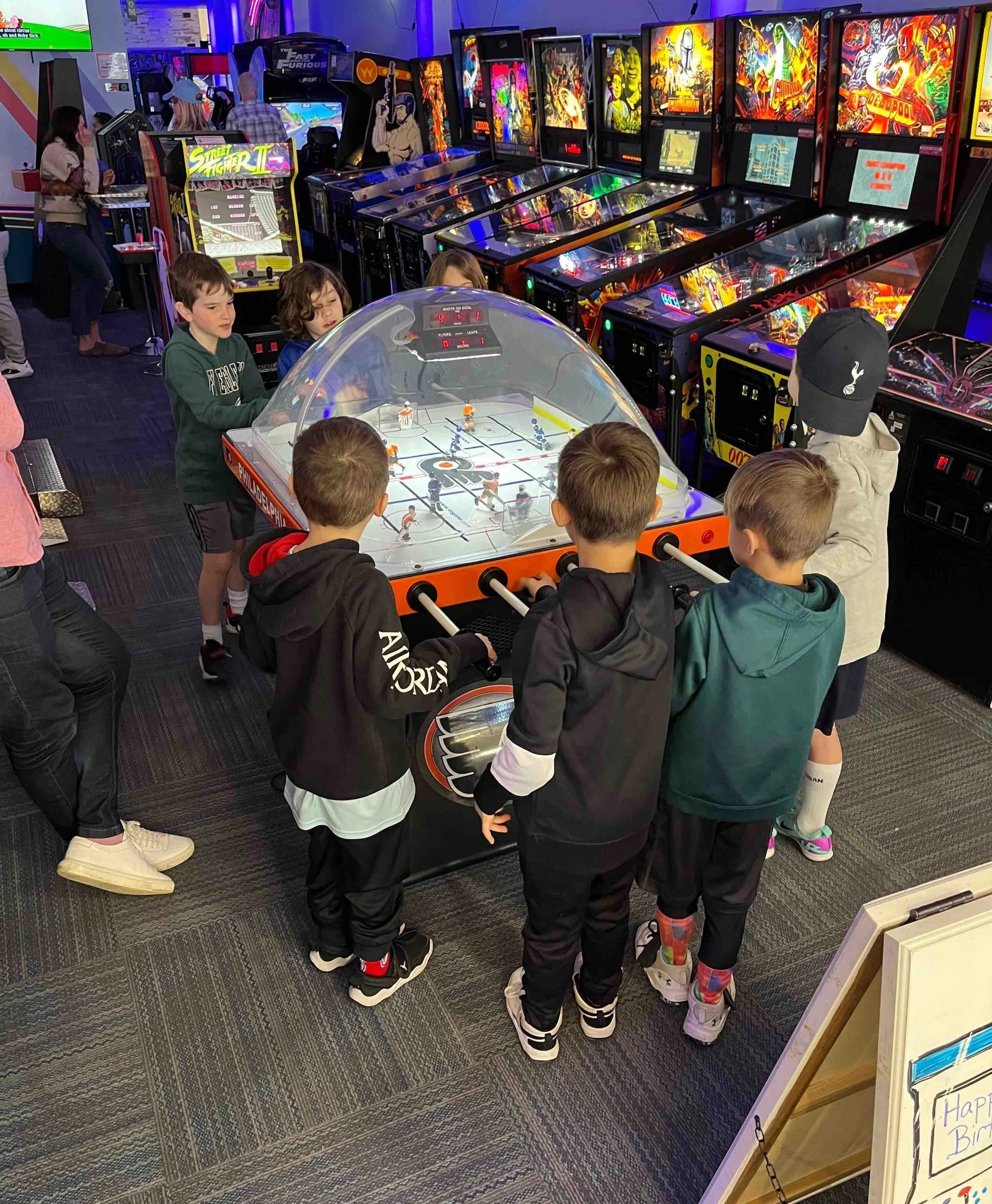 A group of children playing air hockey at an arcade surrounded by various arcade games with colorful lights.