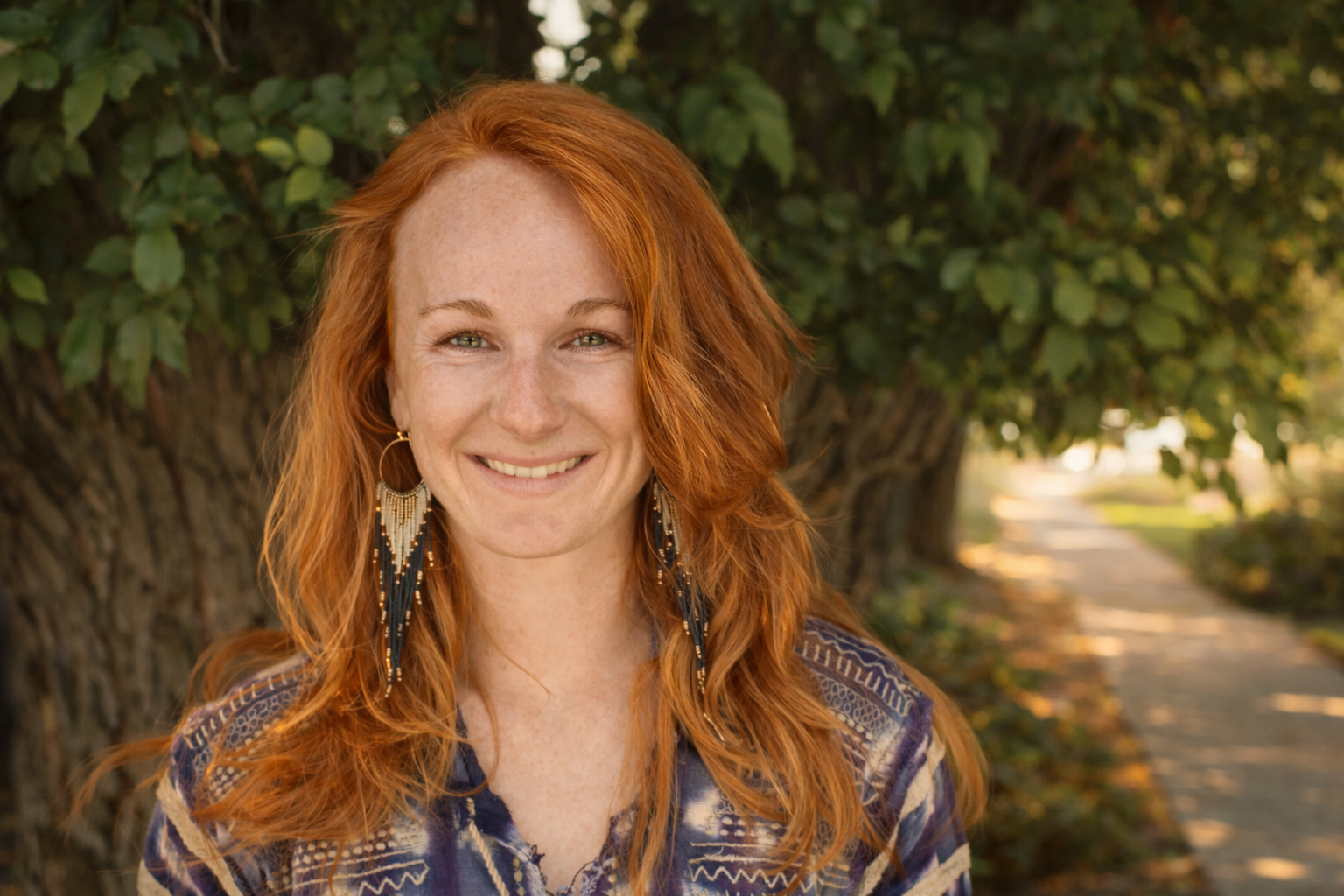 A woman with red hair and earrings smiling outdoors near a tree on a sidewalk.
