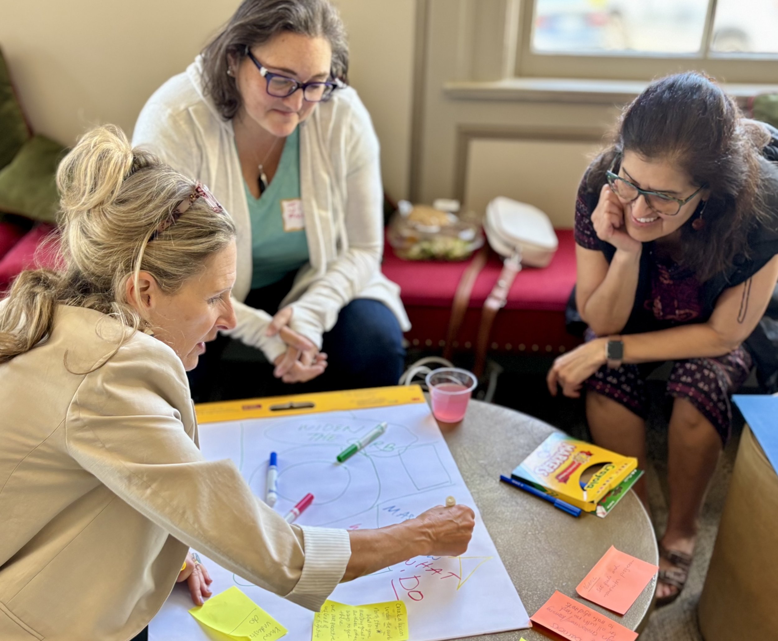 Three women are gathered around a table with colorful markers and a large sheet of paper, engaging in a brainstorming or planning session, with one woman actively writing or drawing on the paper while the other two women look on and smile, in a well-lit room with windows, a bench, and snacks in the background.