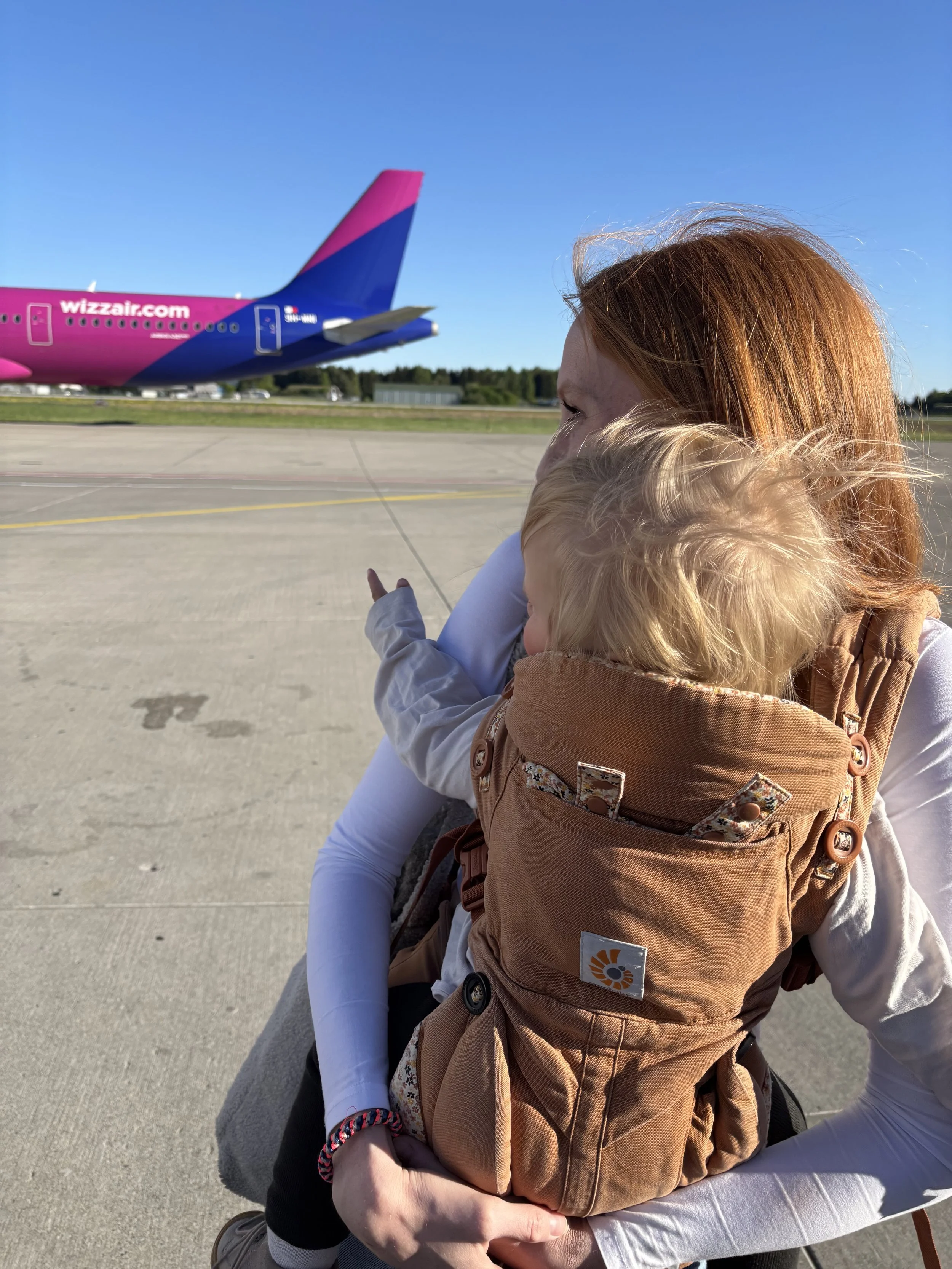 Ein Erwachsener hält ein Kind mit einem Tragesystem, während sie auf einem Flughafen stehen. Im Hintergrund ist ein pink-blaues Flugzeug der Fluggesellschaft Wizz Air auf dem Rollfeld zu sehen.