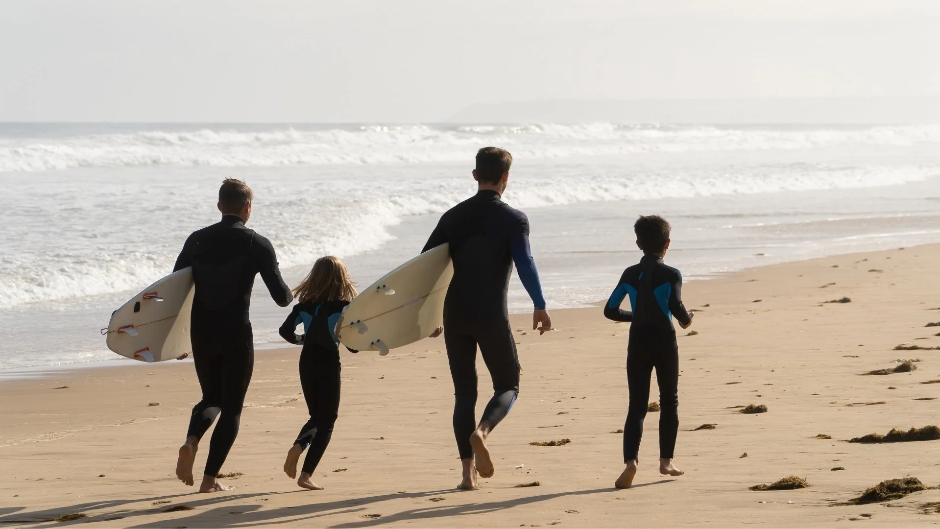 Vier Menschen, zwei Erwachsene und zwei Kinder, zum Surfen in Neoprenanzügen, gehen am Strand entlang, mit Surfbrettern unter dem Arm. Der Strand ist sandig, die Wellen brechen im Meer im Hintergrund.