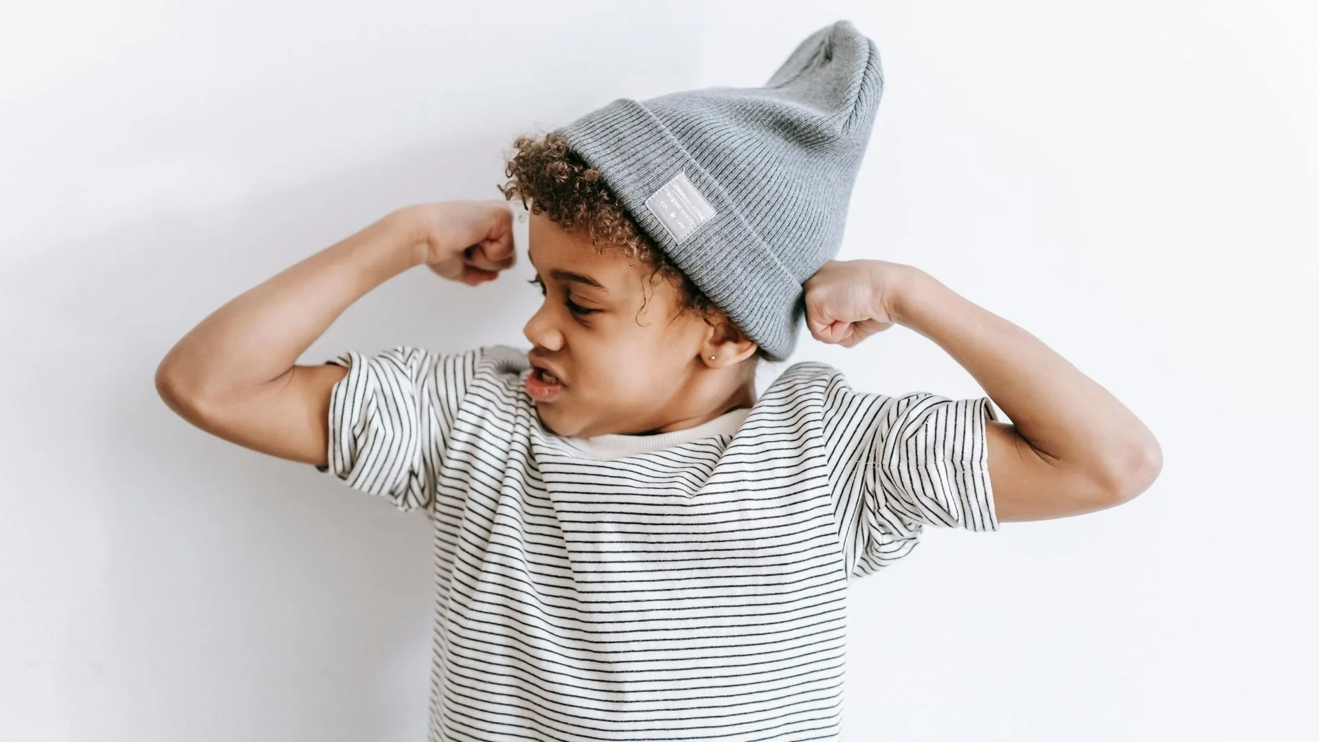 Junge Person trägt grauen Beanie und gestreiftes T-Shirt, macht Muskeln und zeigt Power-Geste.
