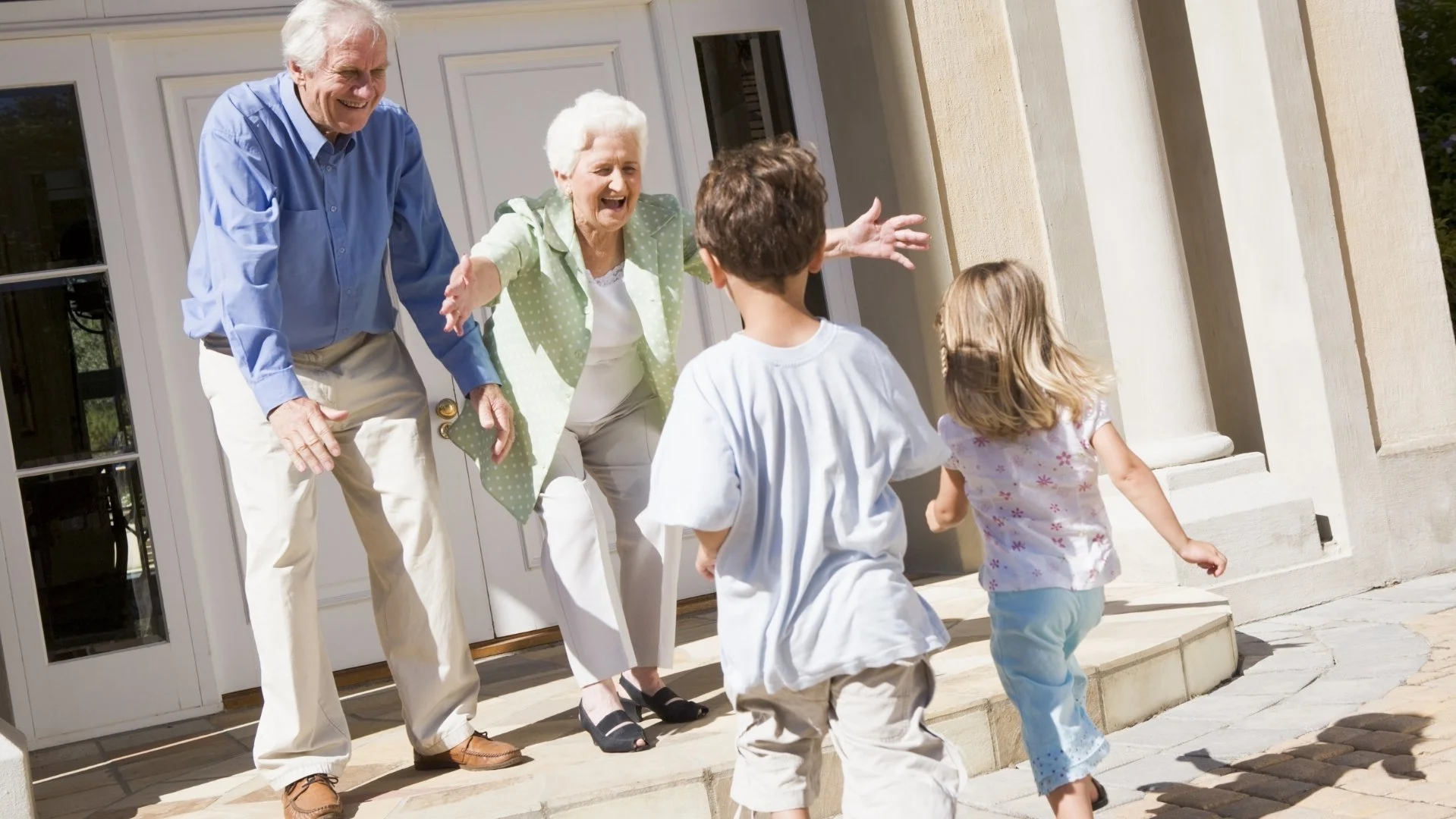 Großeltern begrüßen lachend zwei Kinder vor dem Hauseingang