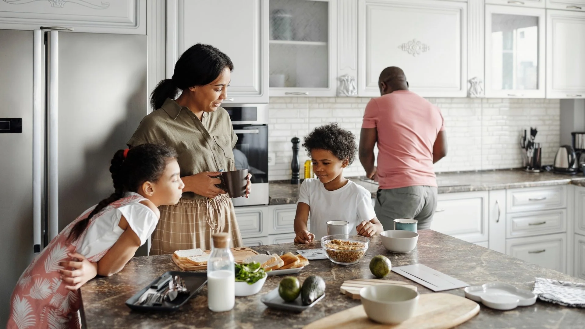 Familie beim Frühstück in der Küche, ein Mann wäscht sich, eine Frau mit Kindern am Tisch, Frühstück auf dem Tisch mit Milch, Brot, Müsli und Obst.