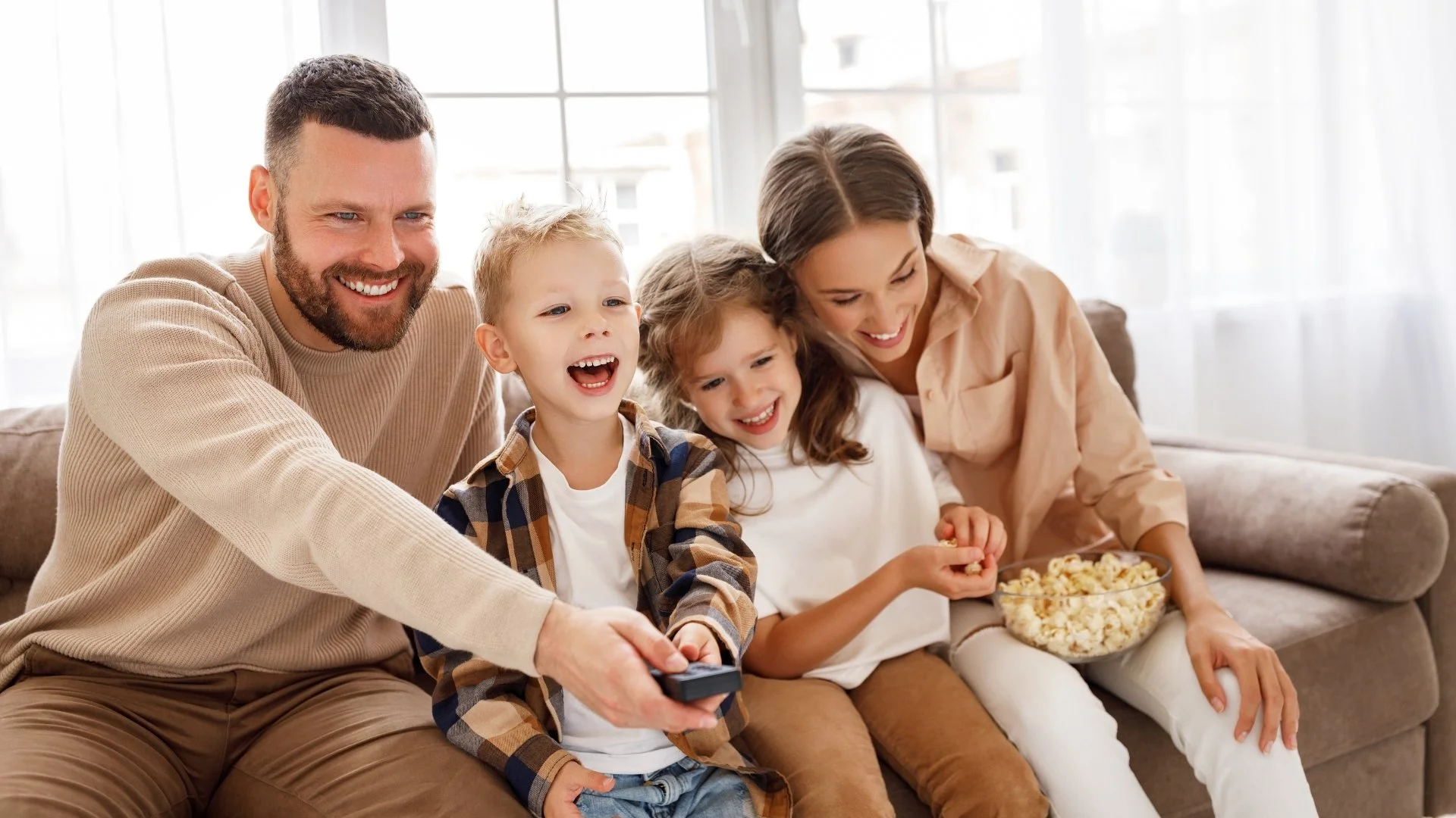 Familie sitzt lachend auf dem Sofa und schaut fernseh. Das Kind im Mittelpunkt hält die Fernbedienung, die anderen essen Popcorn.