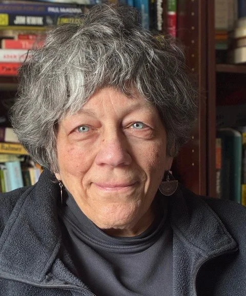 Close-up of an older woman with curly gray hair and blue eyes, wearing a dark jacket and earrings, standing in front of a bookshelf filled with various books.