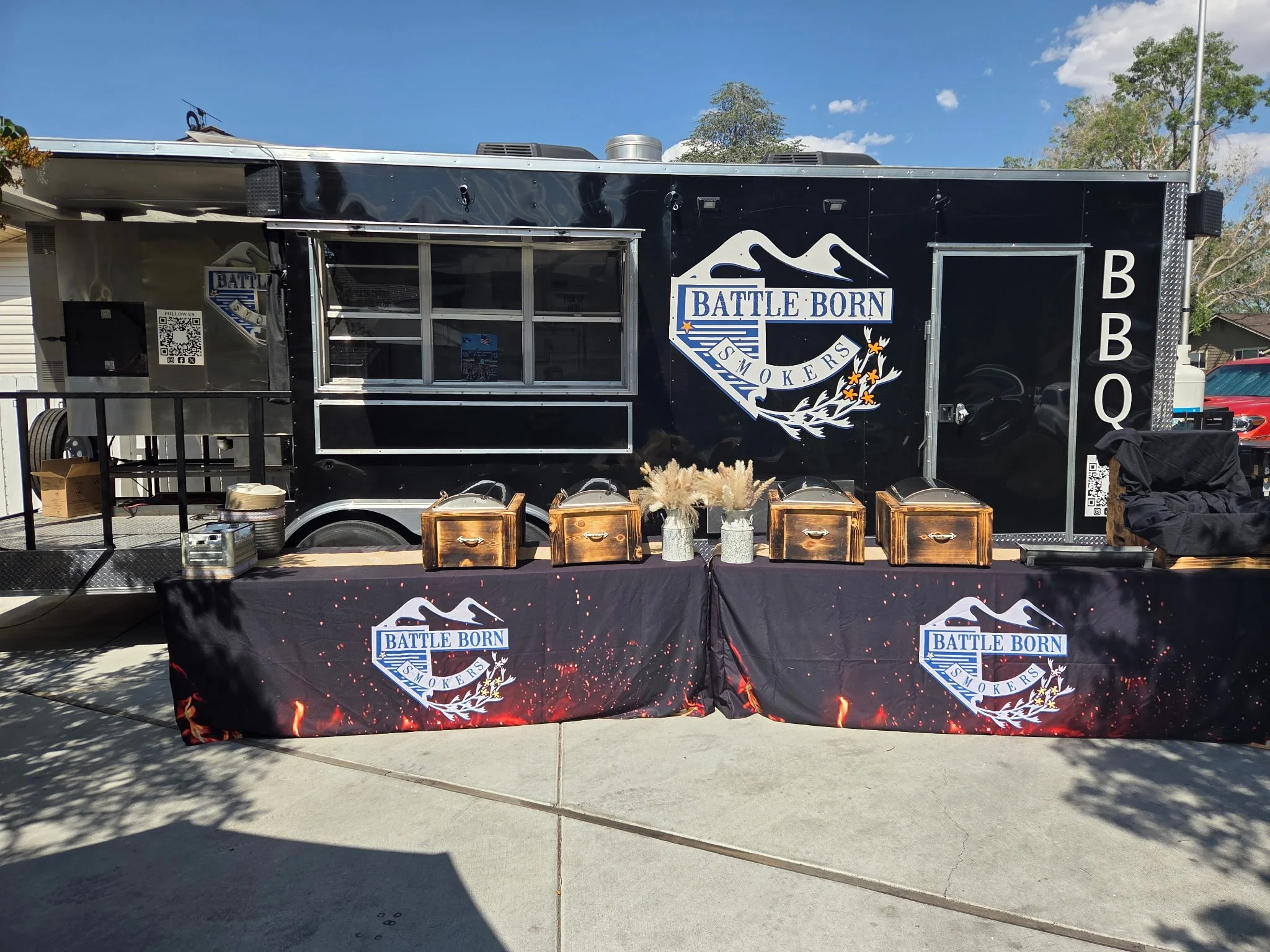 Black BBQ food truck with 'Battle Born Smokers' logo, outdoor seating with tables and decorative vases, and a black tablecloth with the same logo in front.