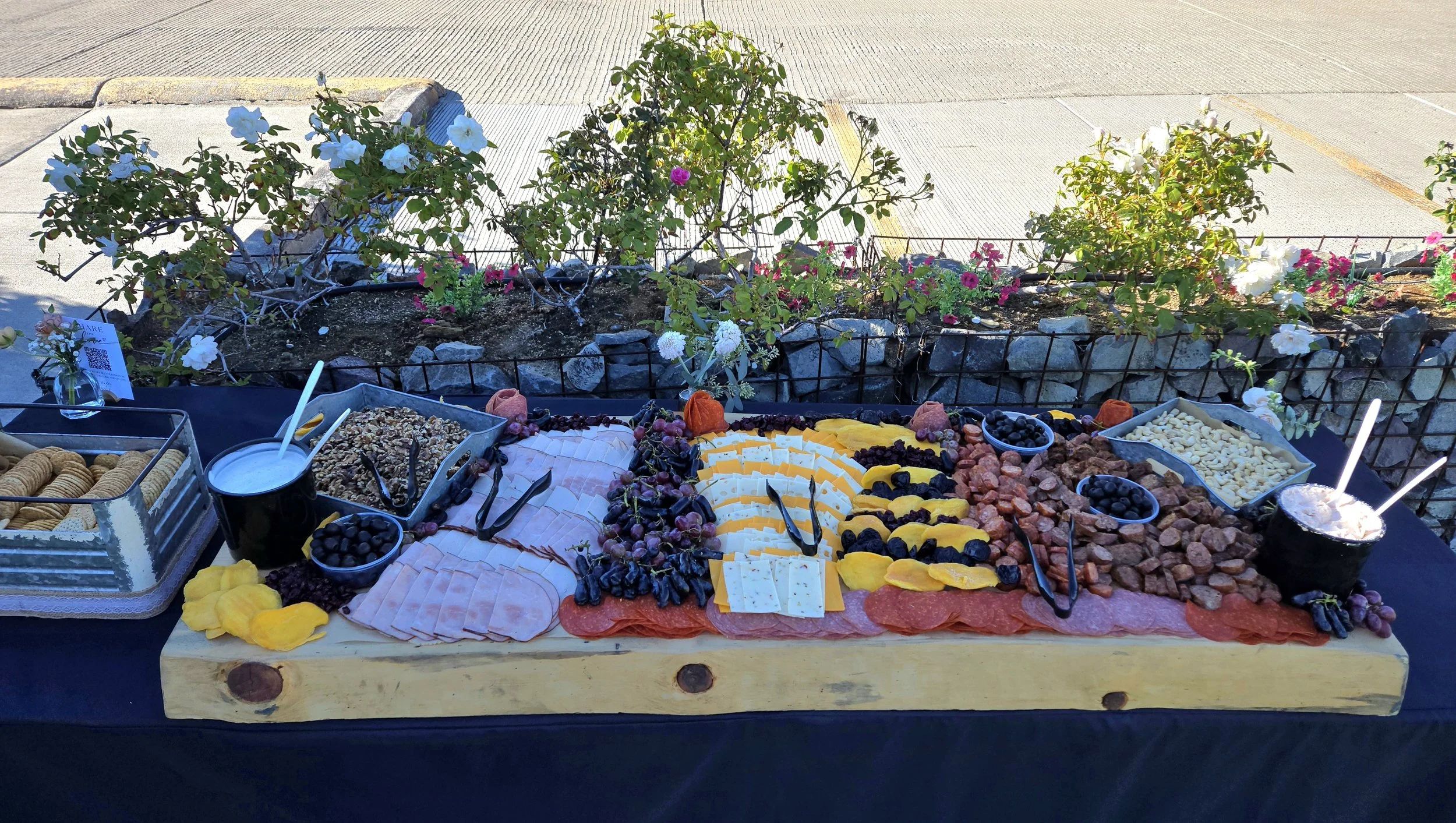 Charcuterie board with various cheeses, meats, grapes, blueberries, crackers, and three cups of drinks on a black table, with white and pink flowers in a garden in the background.