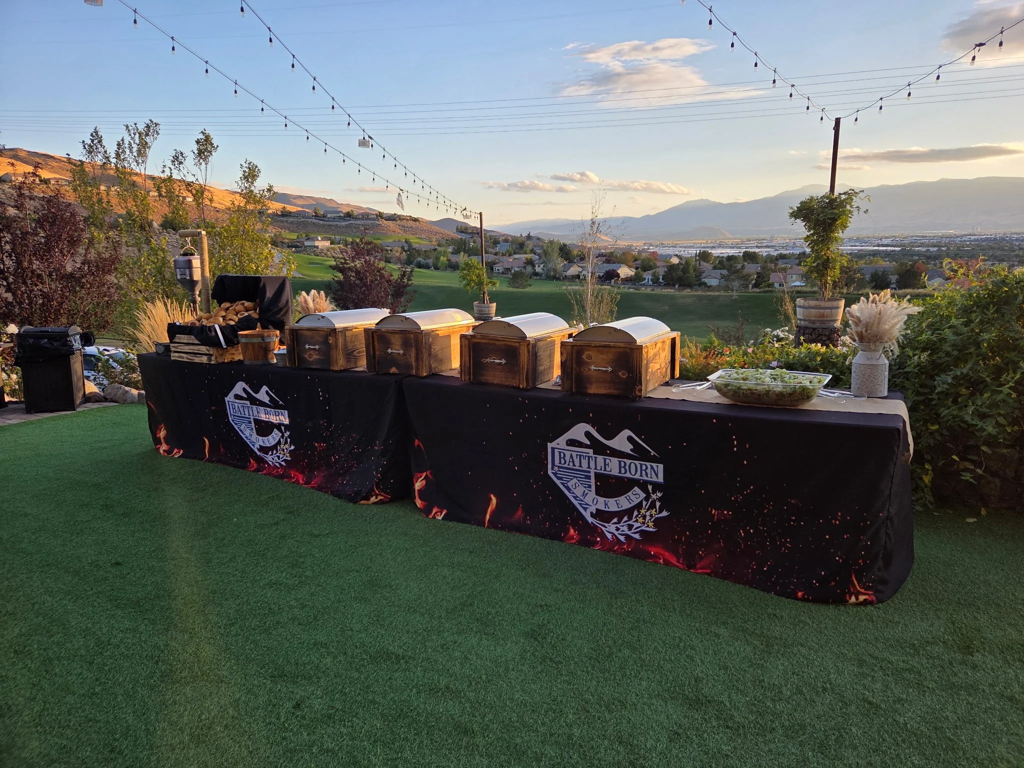 Outdoor buffet table with chafing dishes, salad bowl, and decorations, set against a scenic landscape with rolling hills and string lights at sunset.