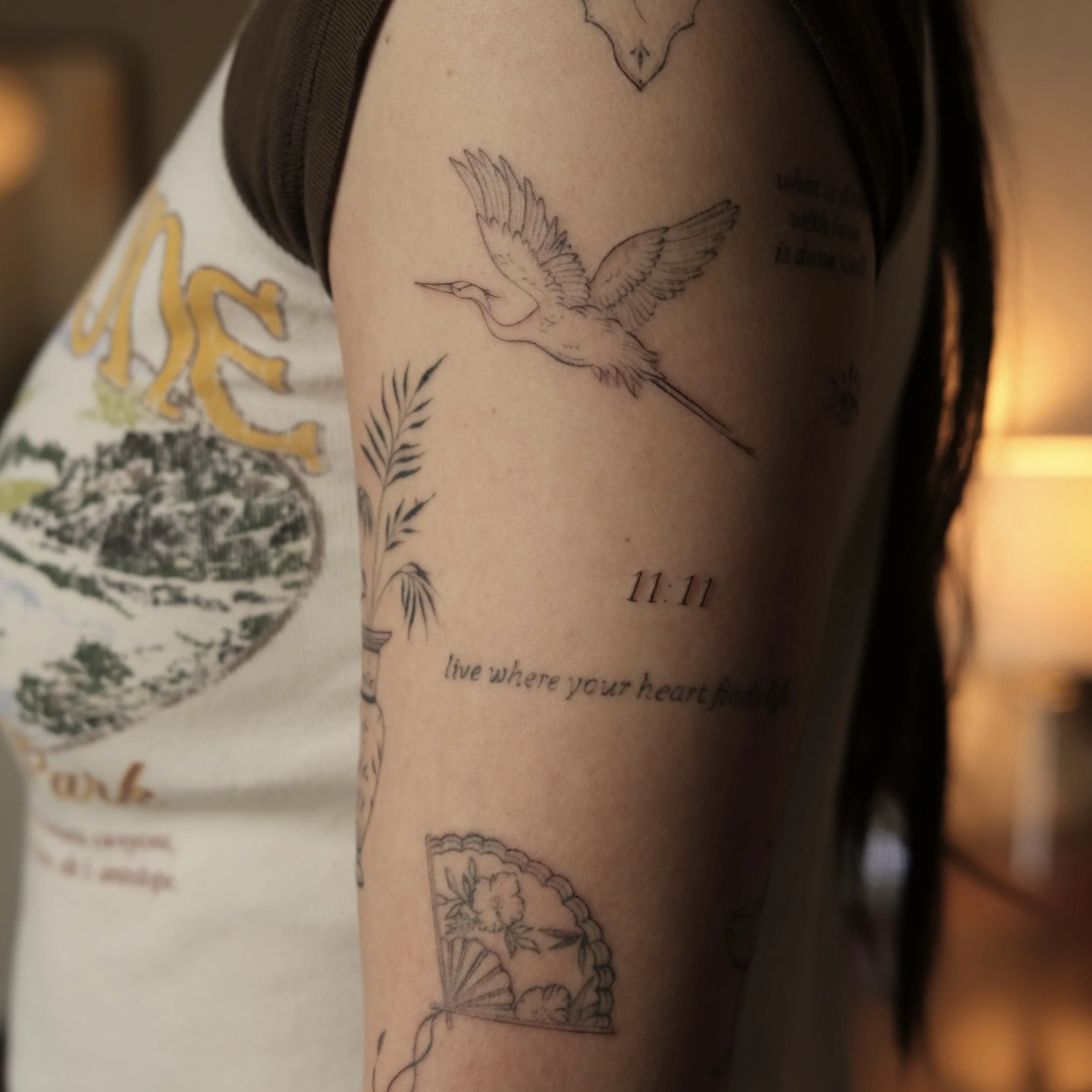 Close-up of a person's upper arm with a black outline tattoo of a hummingbird in flight, surrounded by flowers, with a quote underneath that reads 'live where your heart finds.' The person is wearing a sleeveless shirt with a graphic design.