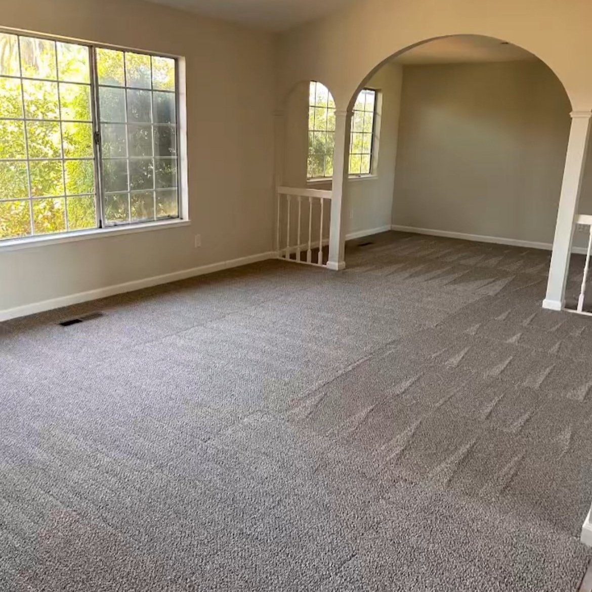 Empty living room with large windows, carpeted floors, and an arched doorway leading to another room.