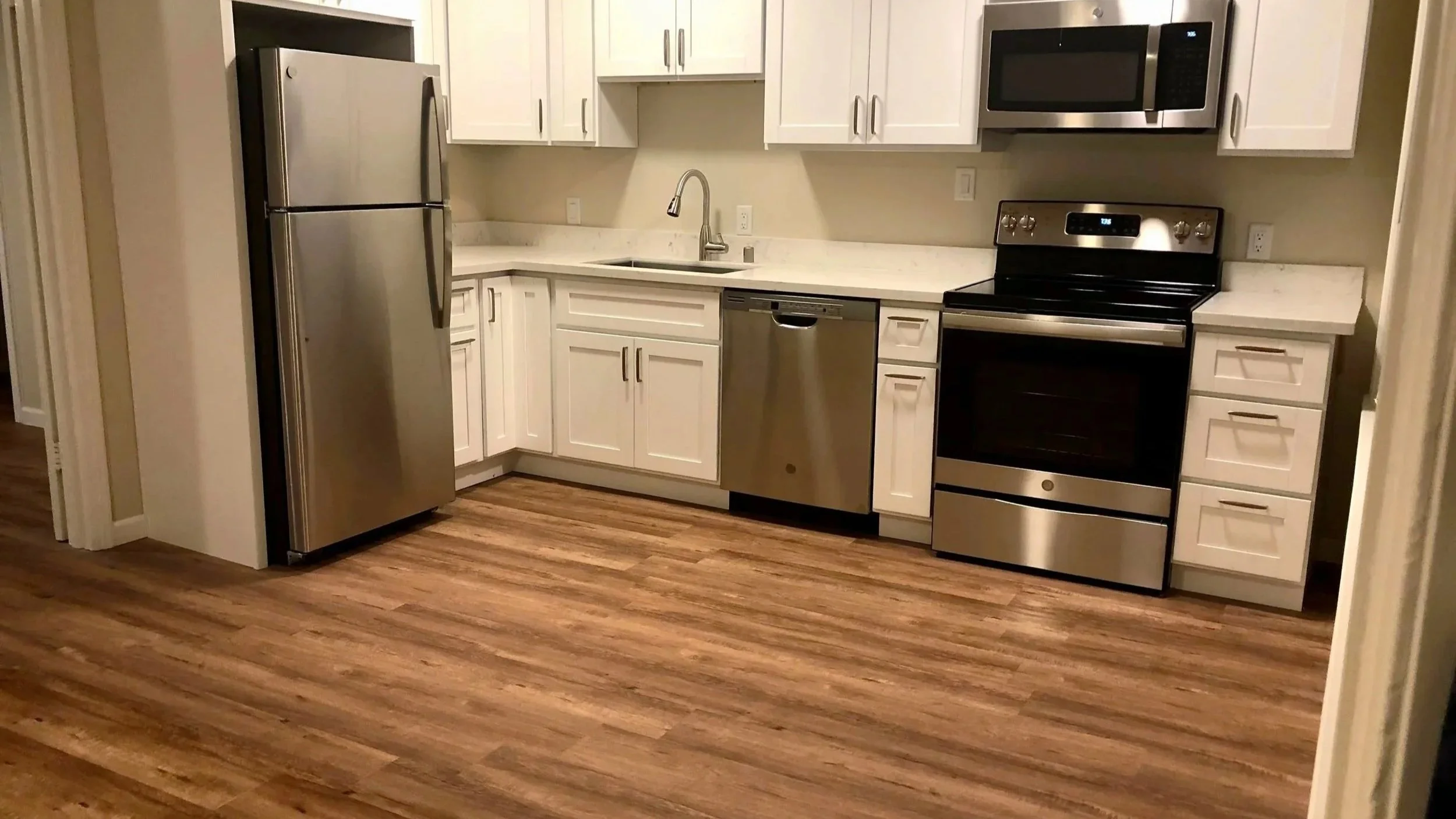 Modern kitchen with white cabinets, stainless steel refrigerator, dishwasher, and oven, along with a microwave, sink, and wood flooring.