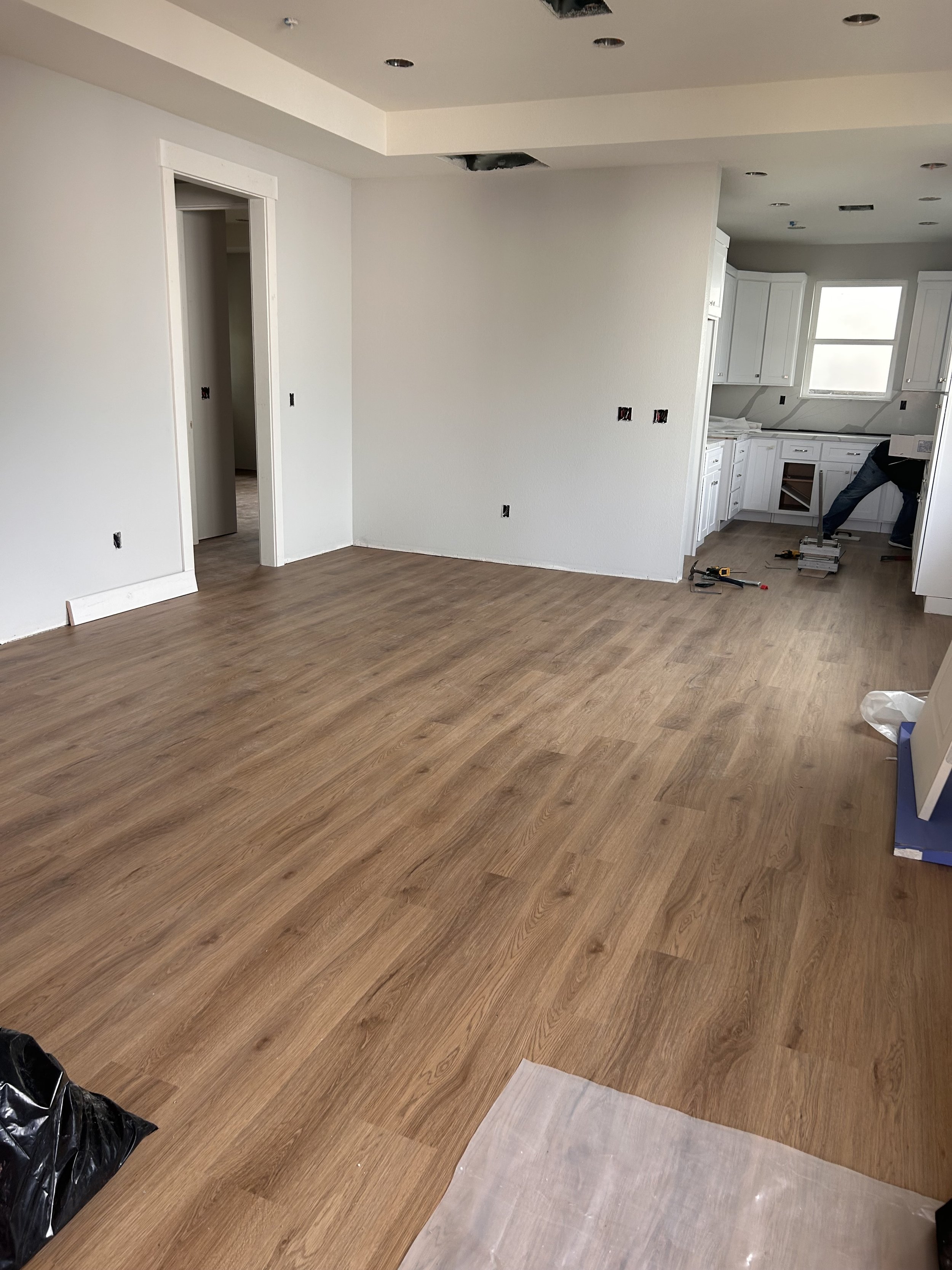 Interior of a room under construction with wood flooring, white walls, and a kitchen area with white cabinets and windows. Tools and plastic sheeting are on the floor, and a worker is partially visible near the kitchen counter.