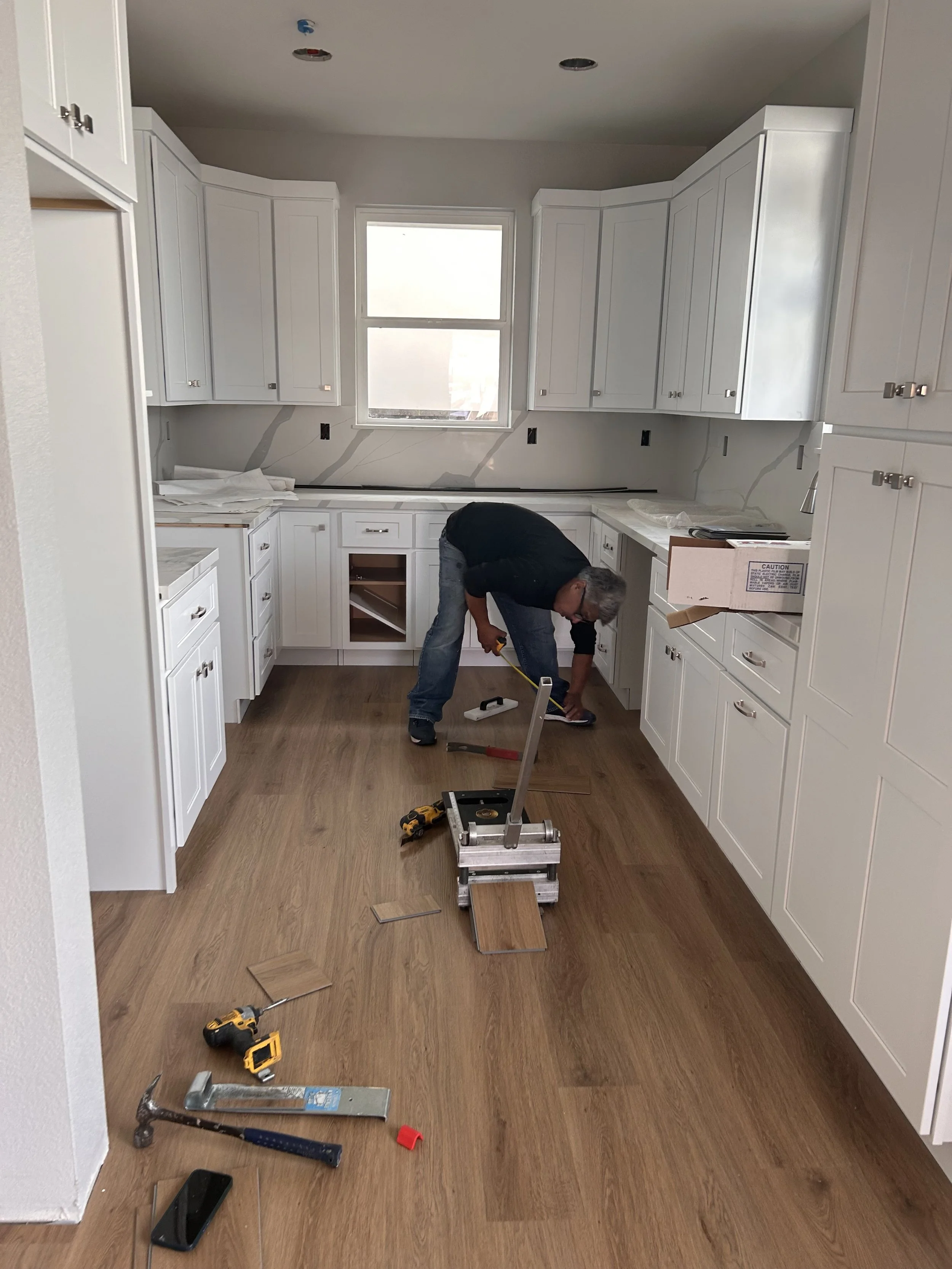 A person working in a kitchen under construction, installing or repairing cabinets. The kitchen has white cabinets, a window, and tools on the floor.