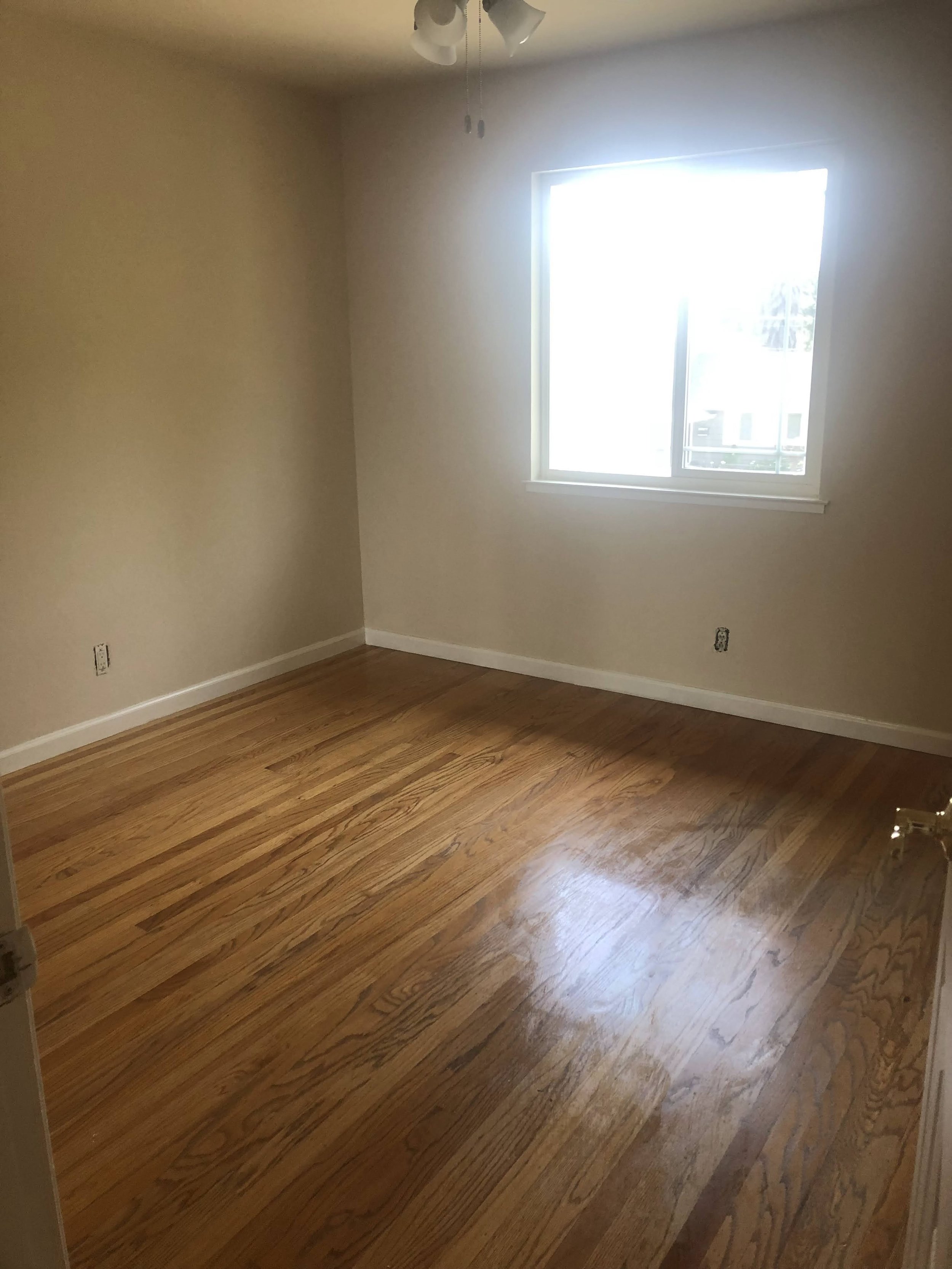 An empty room with hardwood flooring, beige walls, a large window, and a ceiling fan with lights.