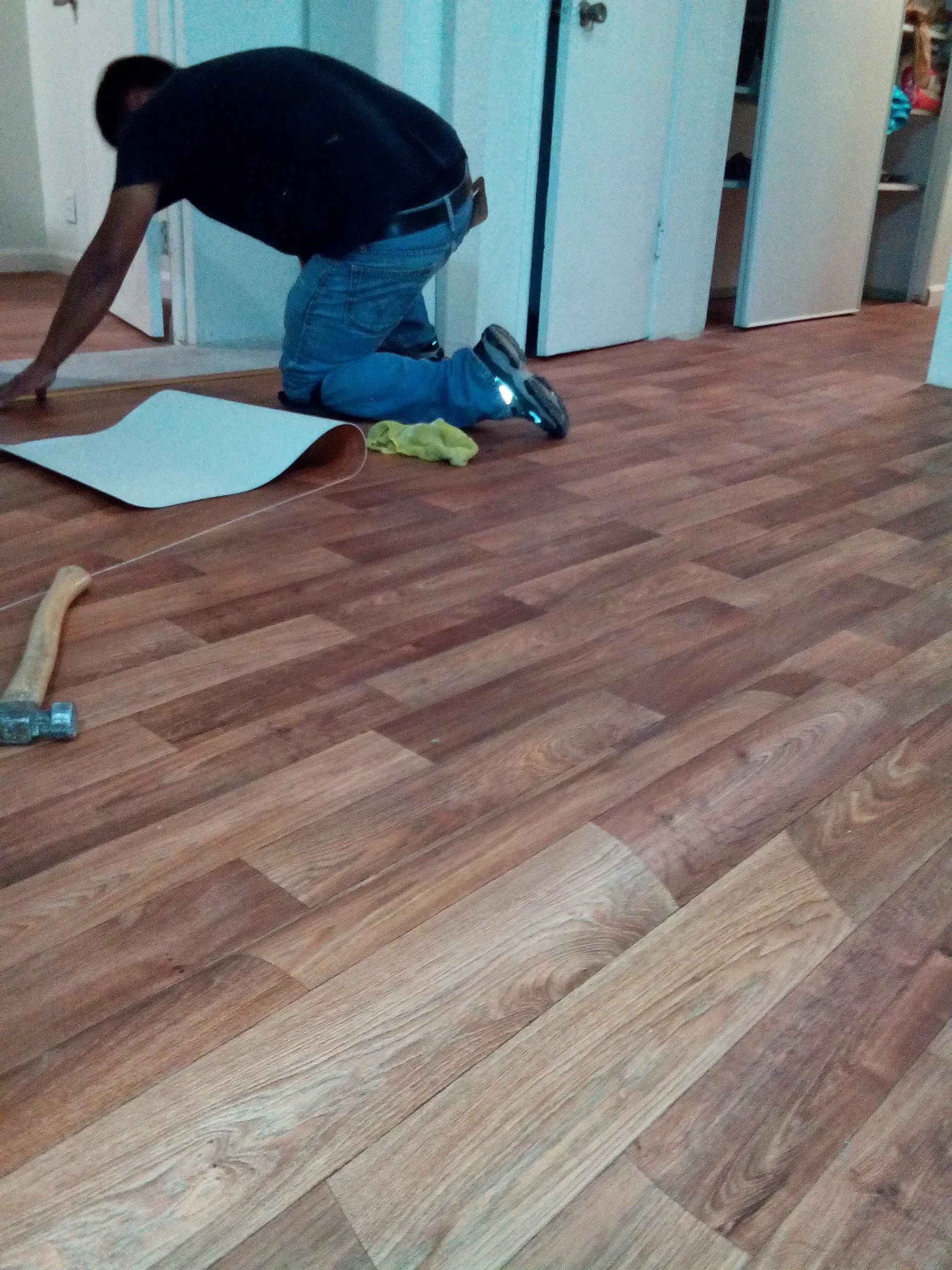 A person kneeling on the wooden floor in front of a closet, cleaning or working on something, with cleaning supplies and tools nearby.