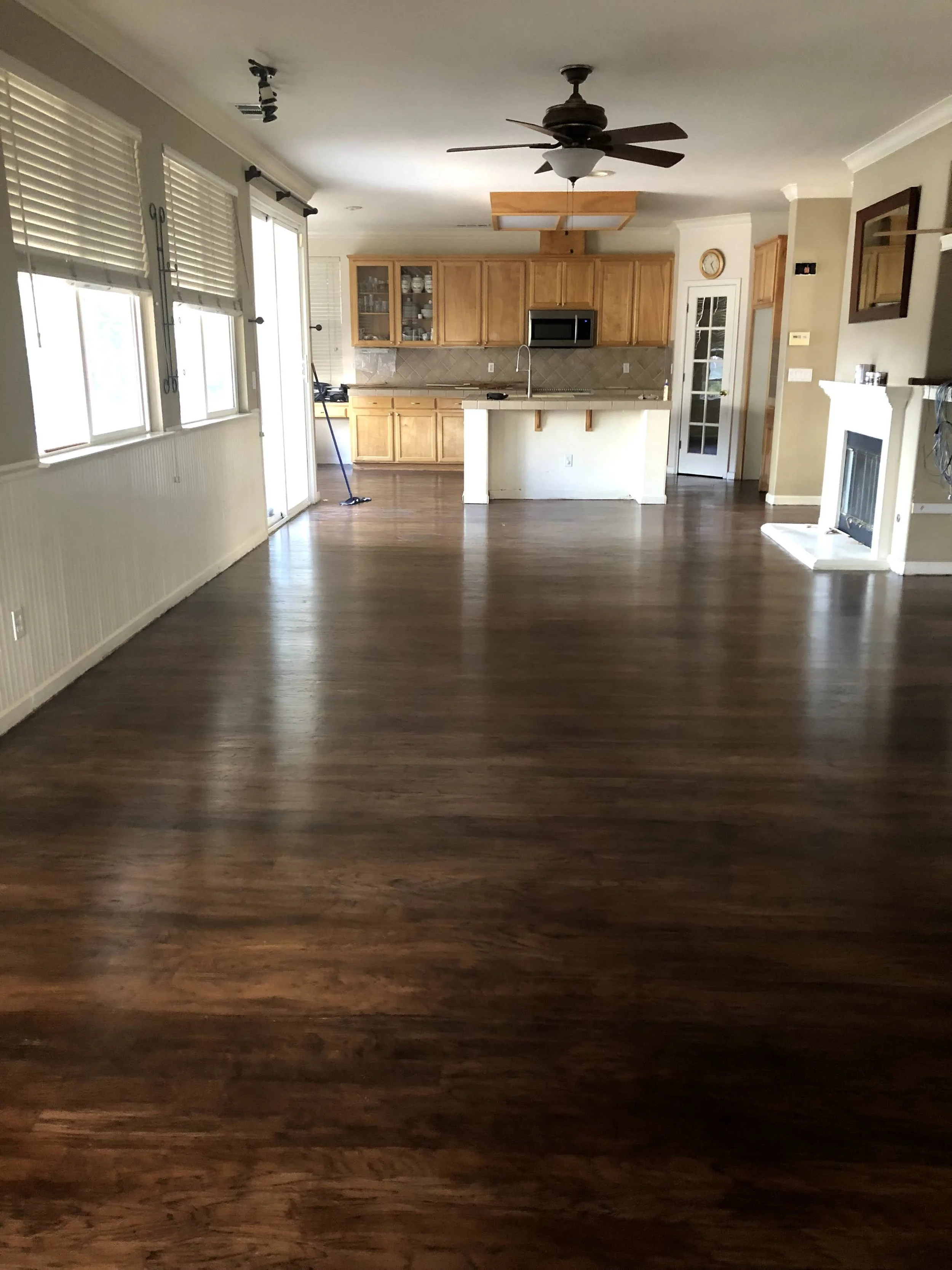 An open-concept living space with hardwood floors, a kitchen with wooden cabinets, a microwave, and a tiled backsplash. Windows with blinds, a ceiling fan, and a fireplace are also visible.