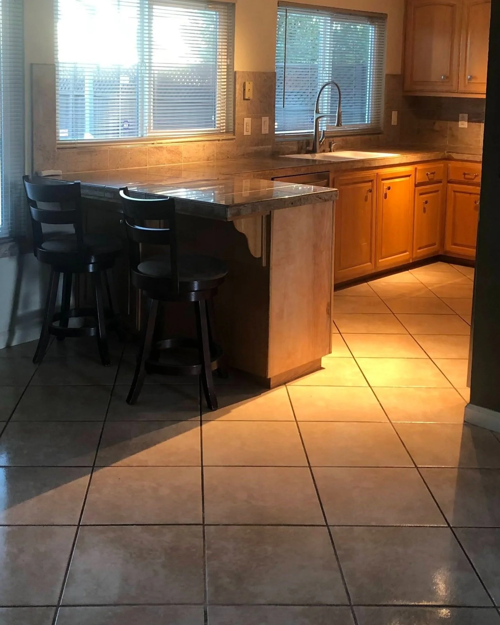 Kitchen with wooden cabinets, a granite countertop, a double sink, and two large windows with blinds letting in natural light. There are three barstools at the counter and ceramic floor tiles.