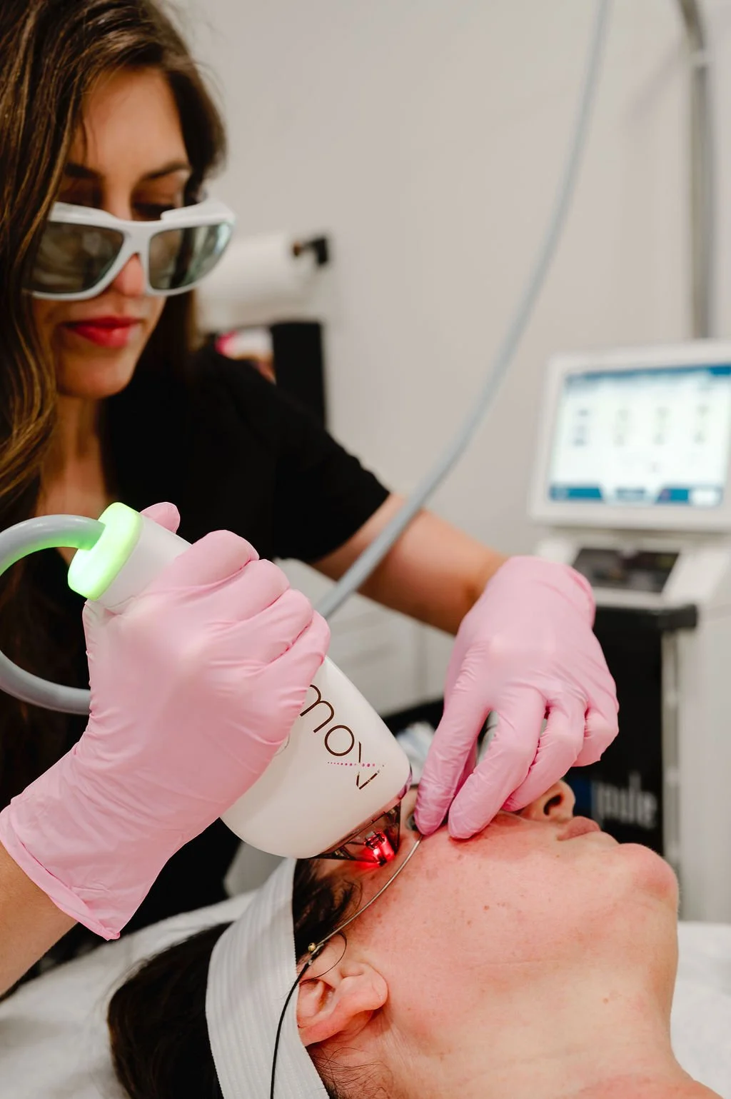 Woman performing facial laser treatment on a client, using a device labeled 'Moxi', in a medical or spa setting. Both individuals wear eye protection; practitioner wears pink gloves.