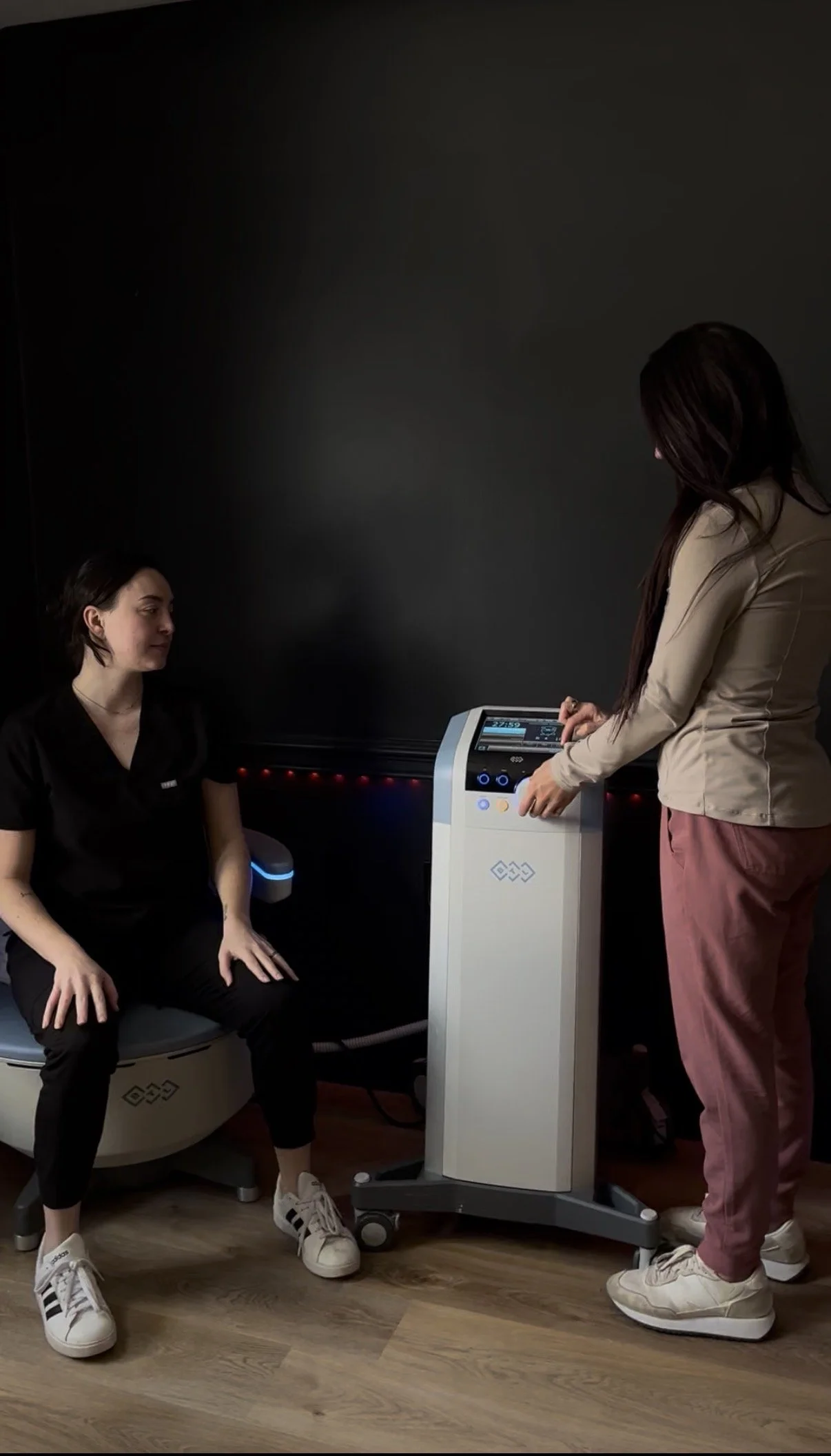 Two women in a medical setting, one sitting on a circular examination chair and the other standing beside a medical device, which is positioned on a mobile stand, against a dark background.